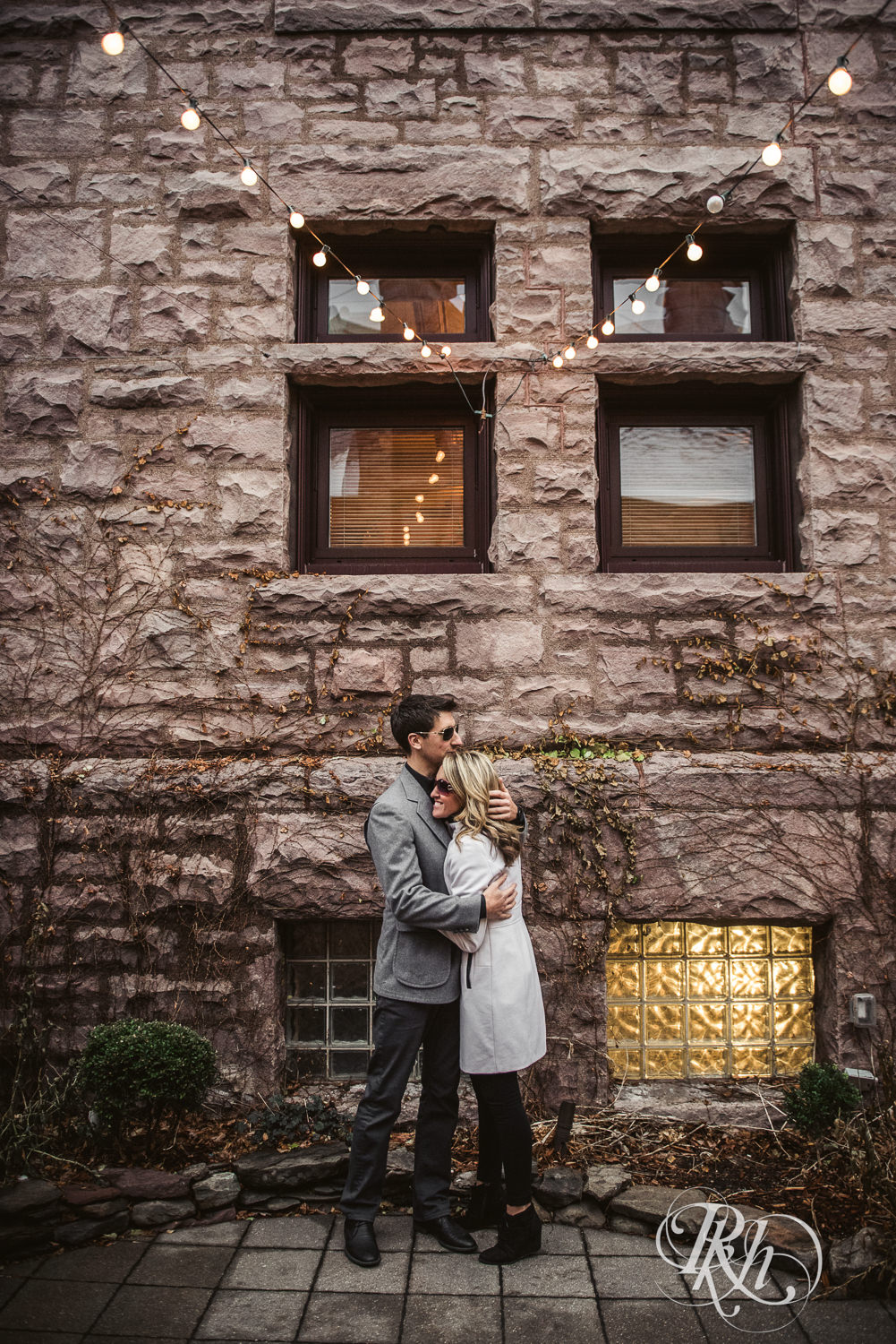 Man and woman in sunglasses and winter coats snuggle in front of the Van Dusen Mansion in Minneapolis, Minnesota.