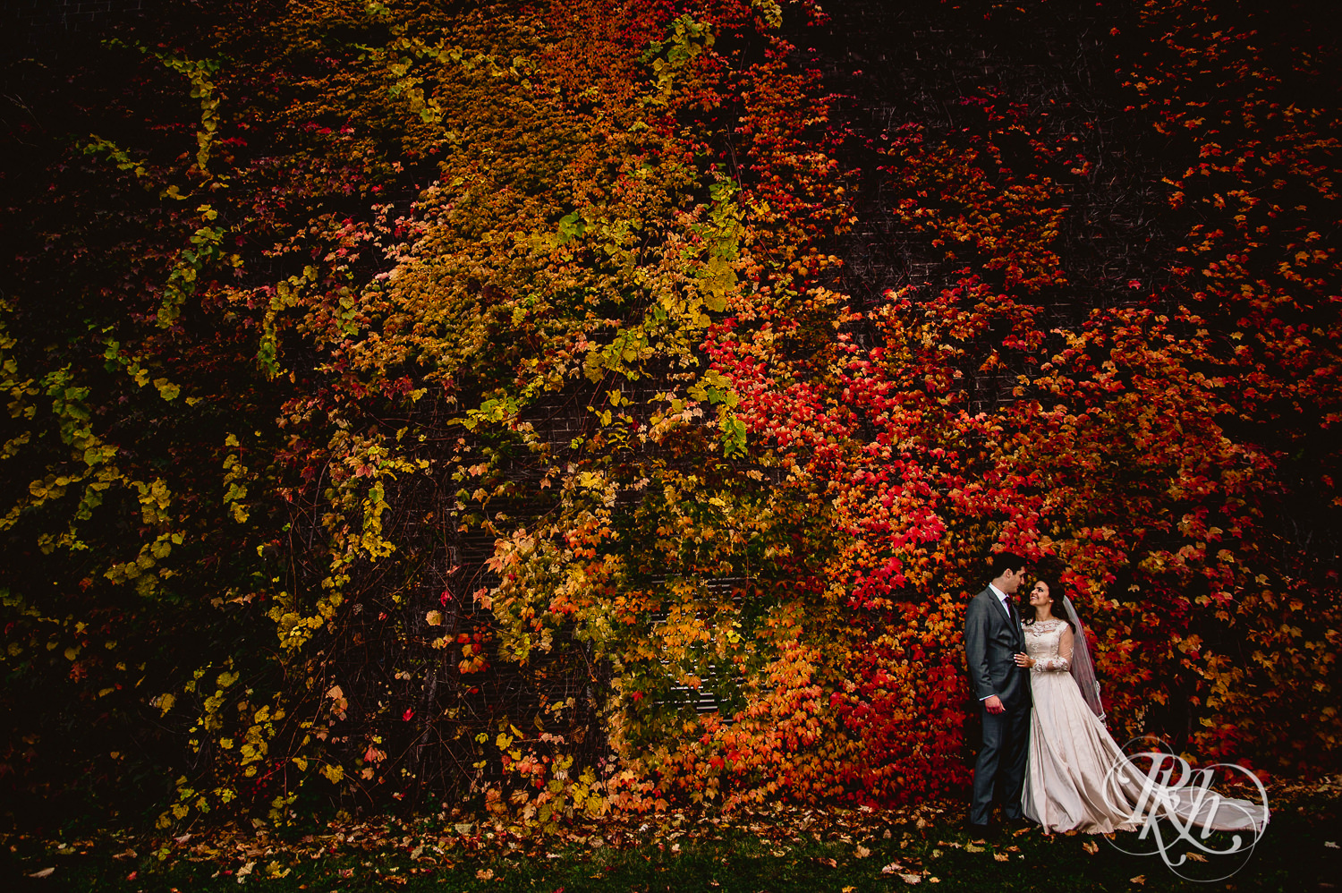 Bride and groom smile in front of a wall covered in red and orange leaves on a rainy wedding day in Minneapolis, Minnesota.