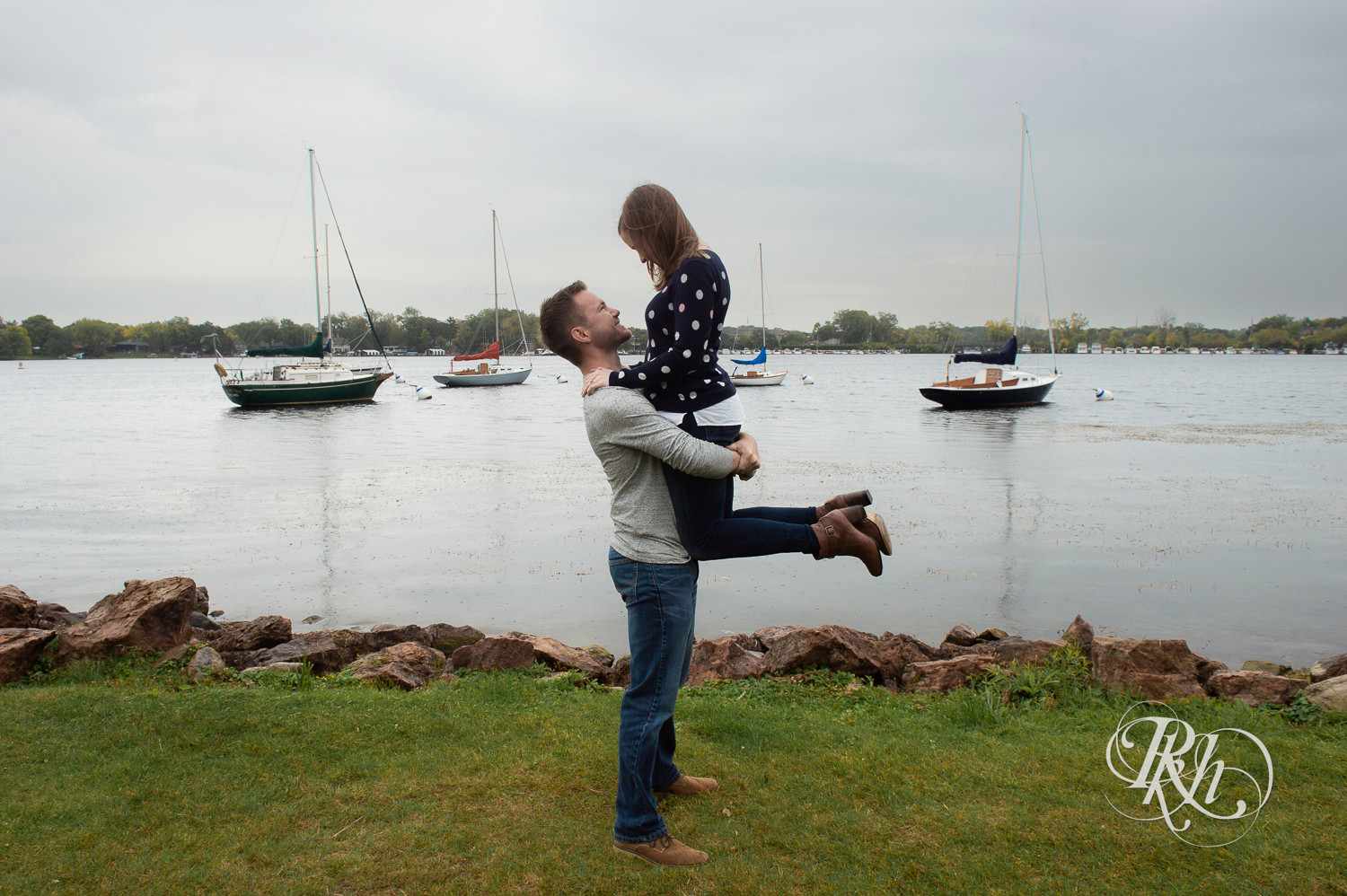 Man and woman in sweaters and jeans smile during rainy day engagement photos in Excelsior, Minnesota.