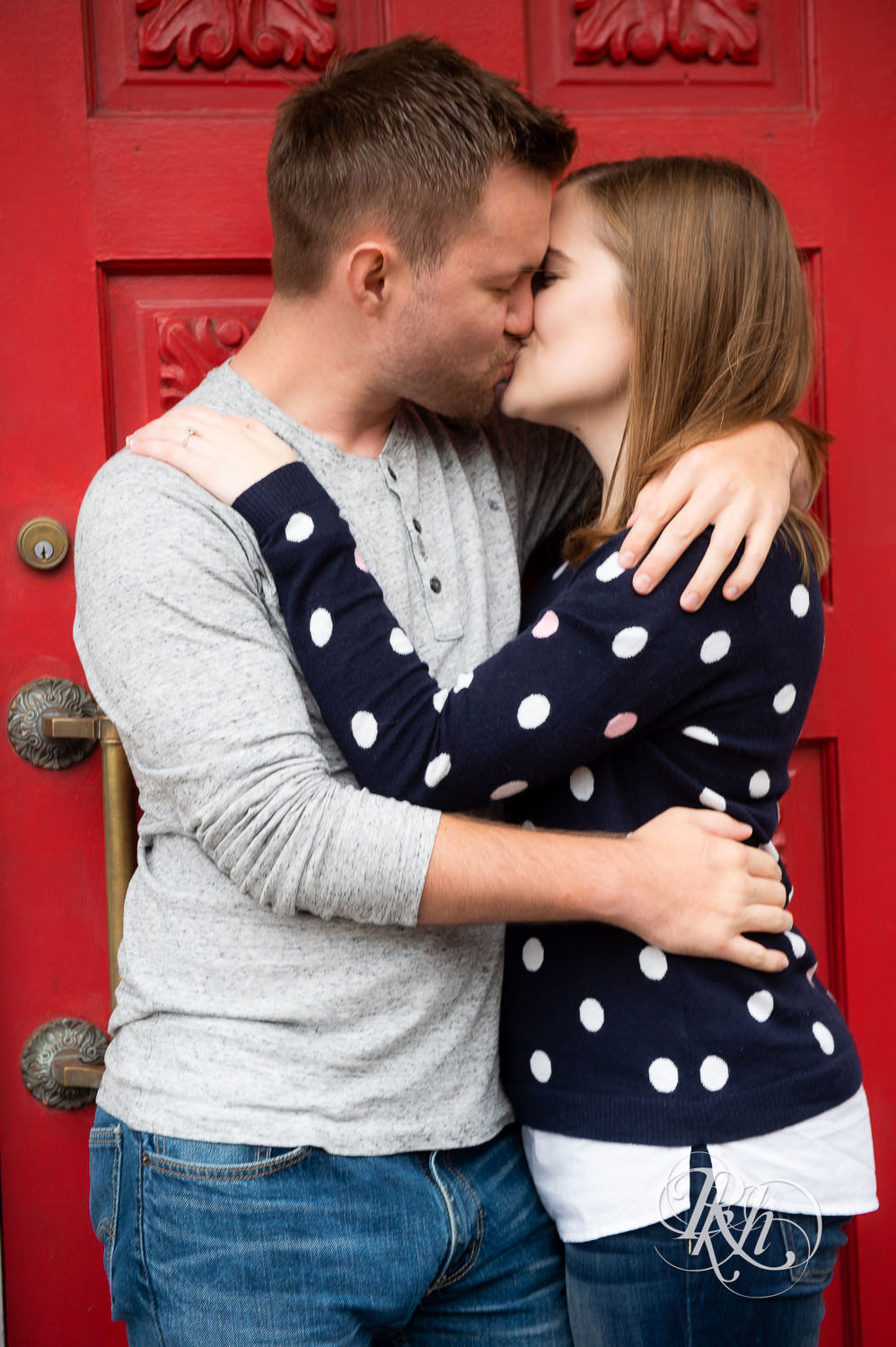 Man and woman kiss against red door on rainy day in Excelsior, Minnesota.