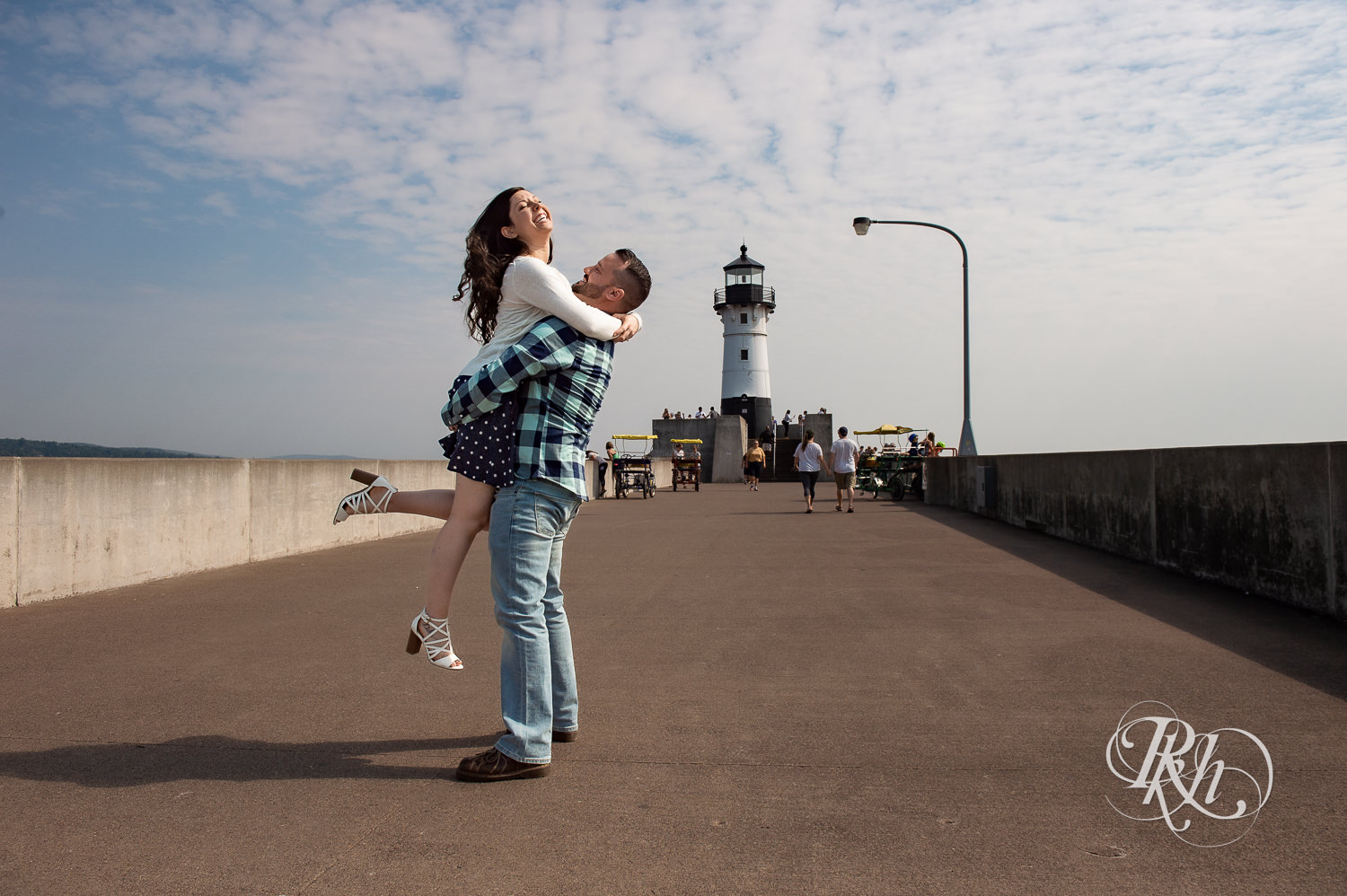 Man and woman laugh with lighthouse in background during Canal Park engagement photos in Duluth, Minnesota.