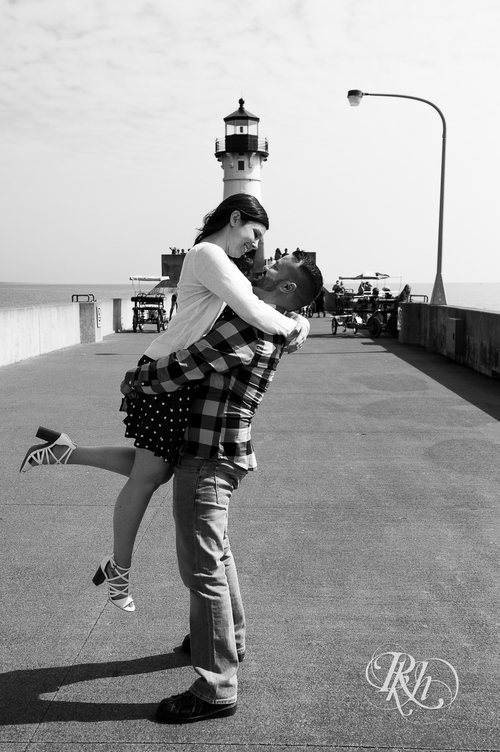 Man and woman laugh with lighthouse in background during Canal Park engagement photos in Duluth, Minnesota.