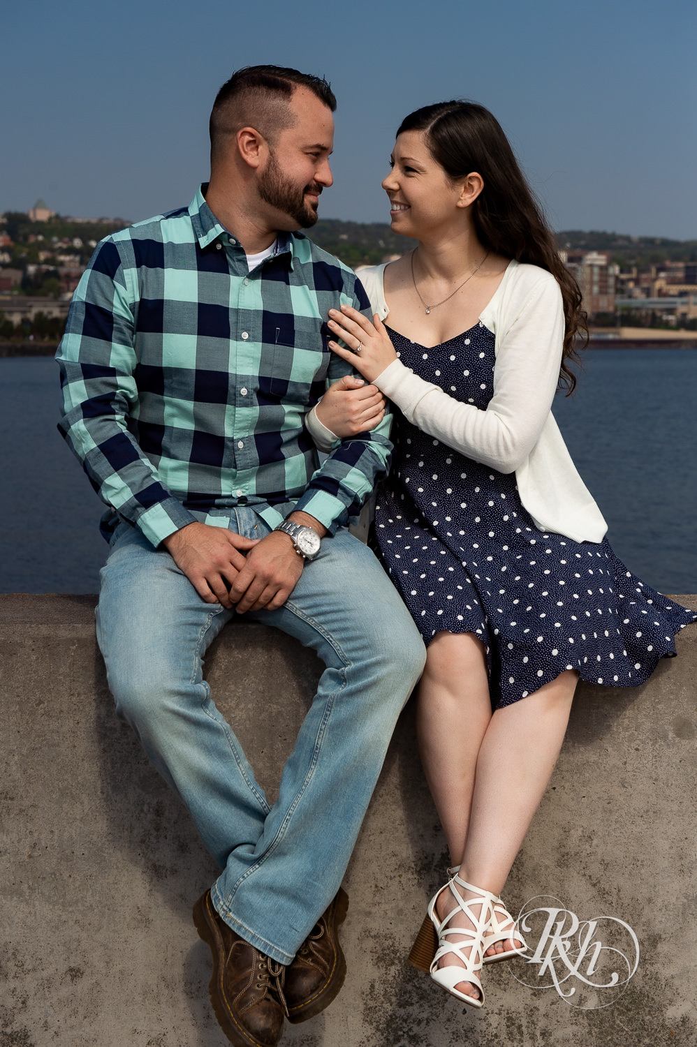 Man and woman smile with city in background during Canal Park engagement photos in Duluth, Minnesota.