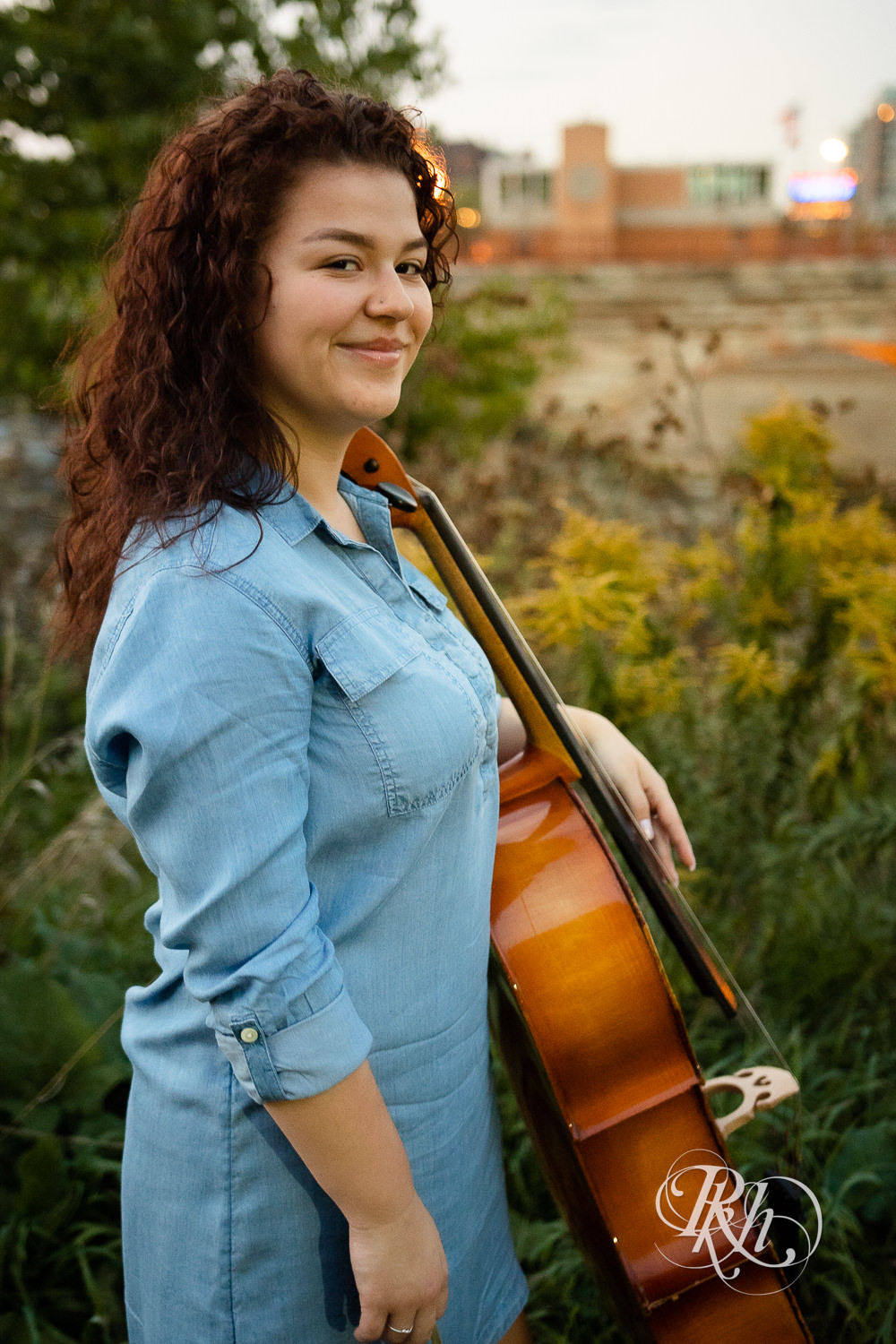 Senior girl in denim dress with curly hair smiles playing cello during sunset in Minneapolis, Minnesota.