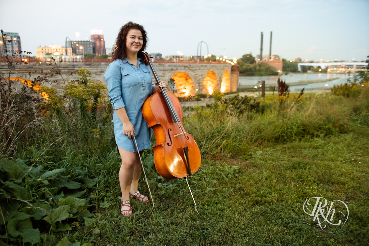 Senior girl in denim dress with curly hair smiles with cello during sunset in Minneapolis, Minnesota.