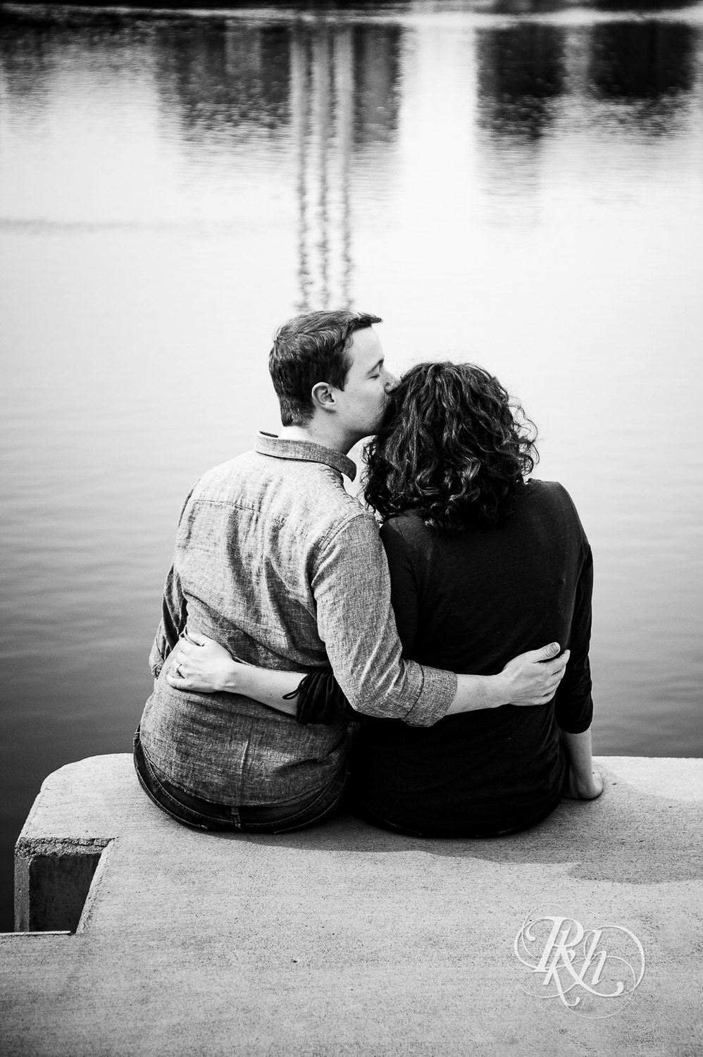 Man and woman kiss in front of the river and Minneapolis skyline during St. Anthony Main engagement photos.