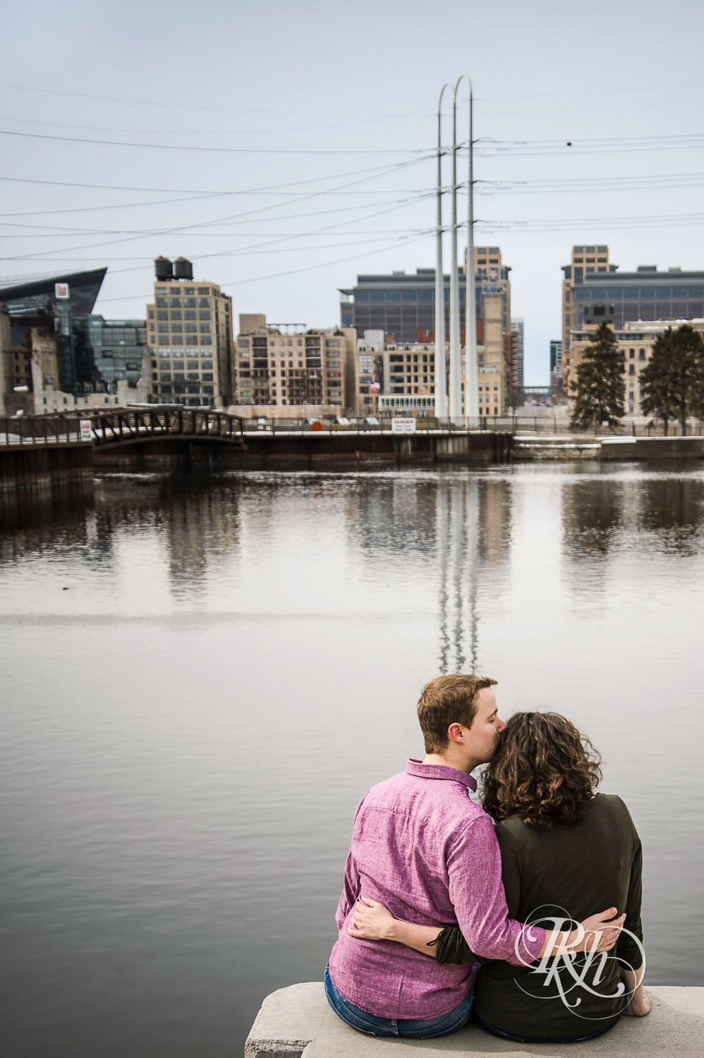 Man and woman smile in front of the river and Minneapolis skyline during St. Anthony Main engagement photos.
