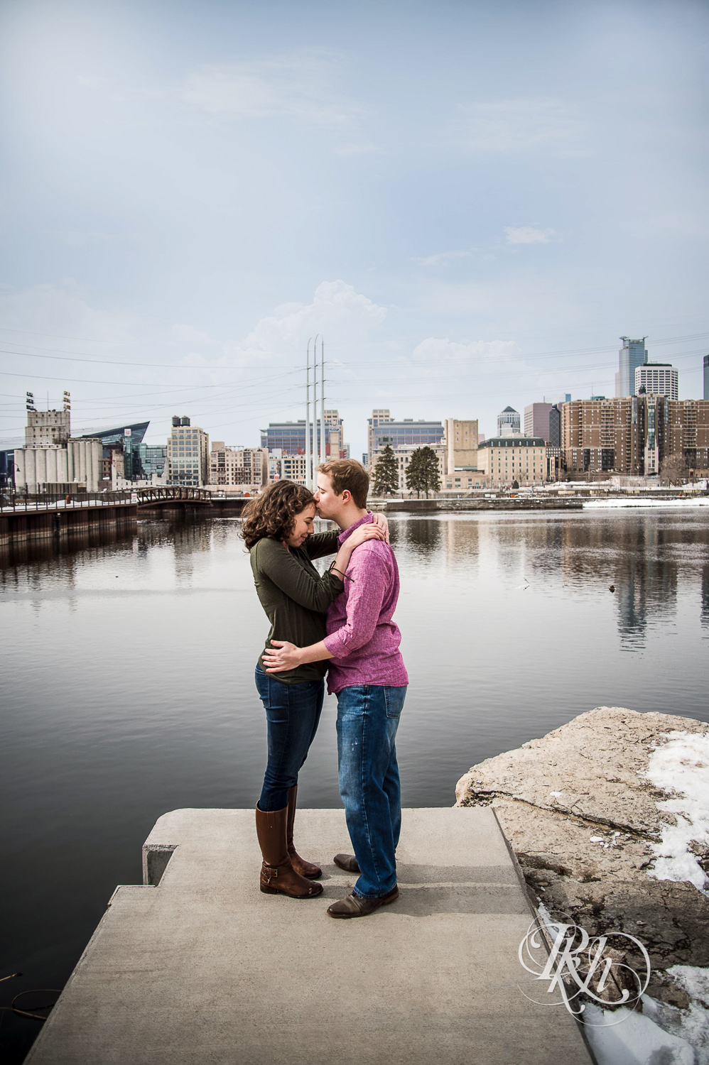Man and woman kiss in front of the river and Minneapolis skyline during St. Anthony Main engagement photos.
