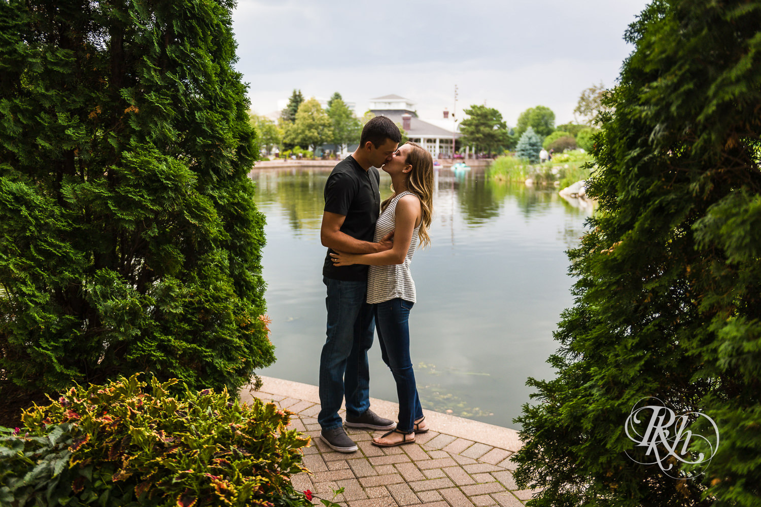 Man and woman in T-shirts and jeans kiss between trees in Centennial Lakes Park in Edina, Minnesota.