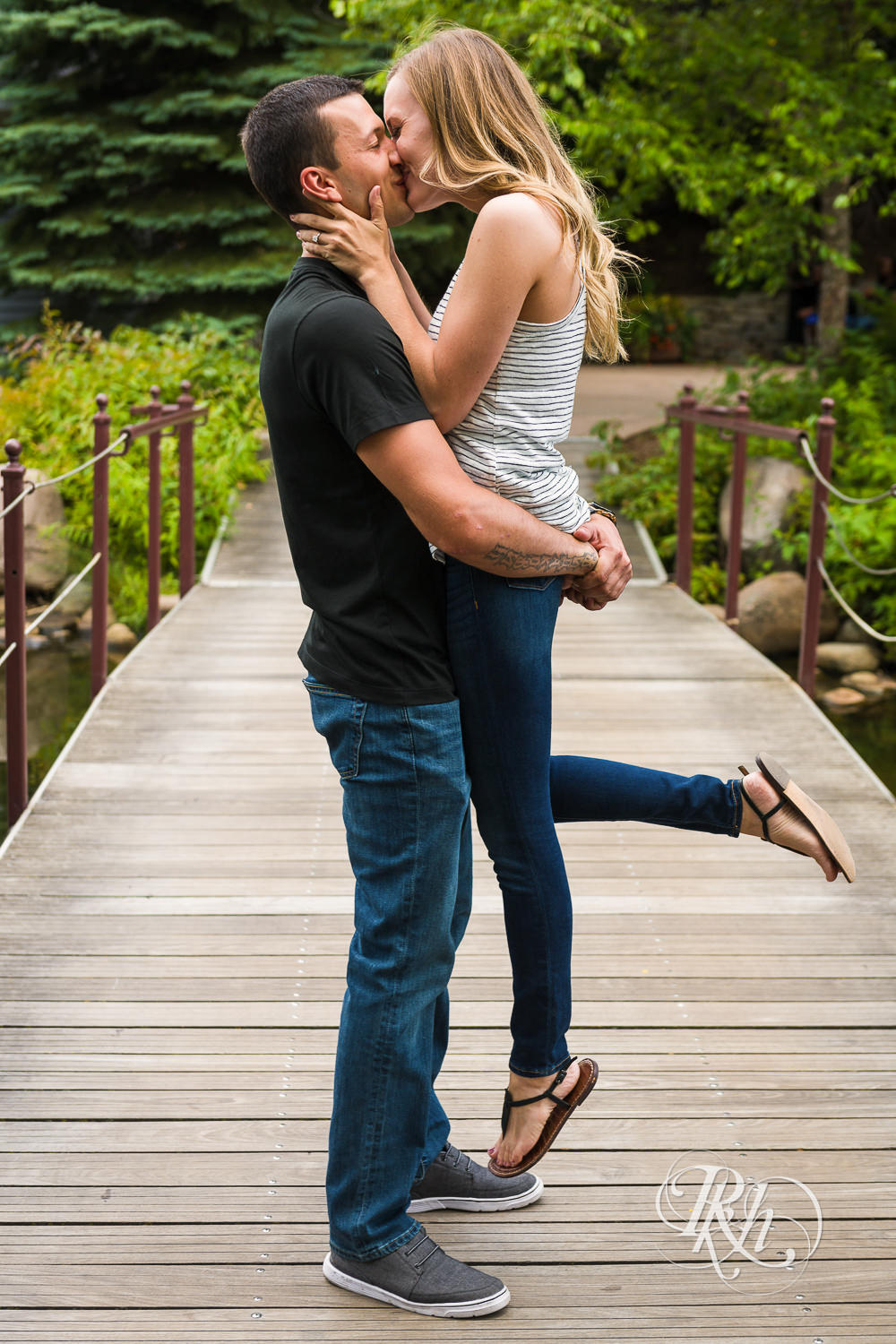 Man and woman in T-shirts and jeans kiss on bridge in Centennial Lakes Park in Edina, Minnesota.