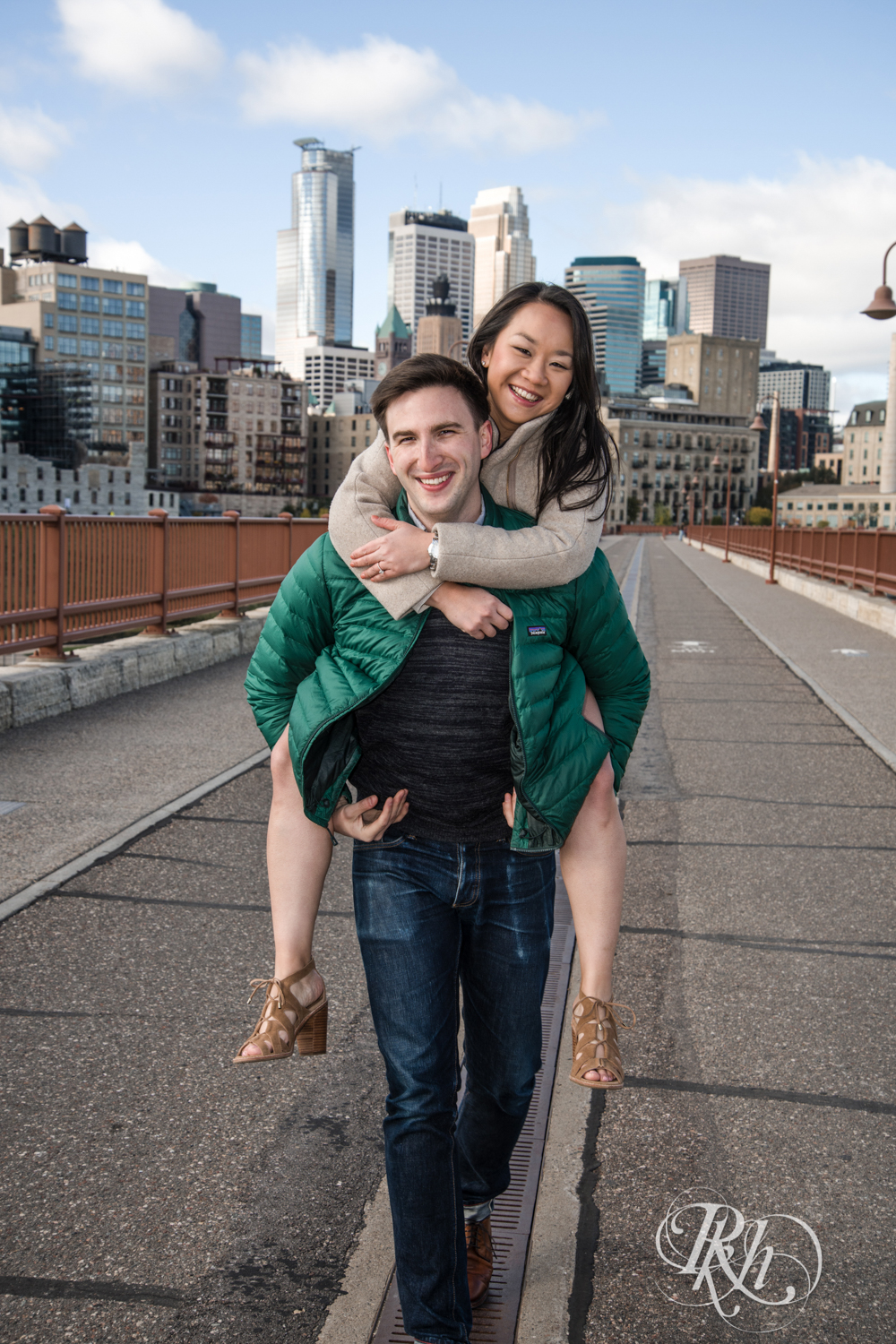 Man in sweater and Asian woman do piggyback ride during Stone Arch Bridge engagement photography in Minneapolis, Minnesota.