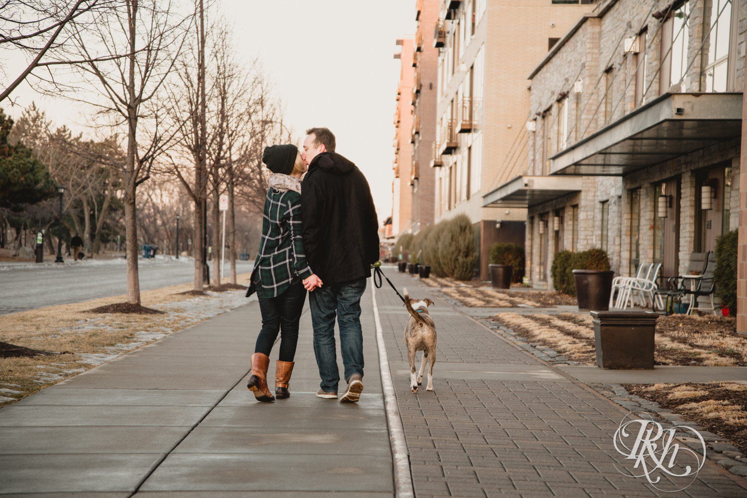 Man and woman kiss while walking their dog during sunrise engagement photography in Minneapolis, Minnesota.