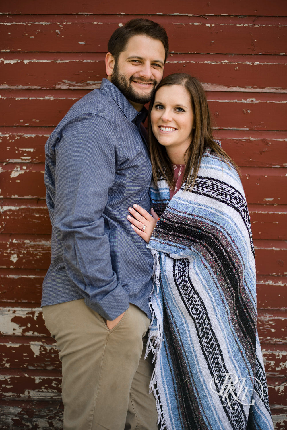 Man and woman smile in front of red barn during engagement photography session at home in Annandale, Minnesota.