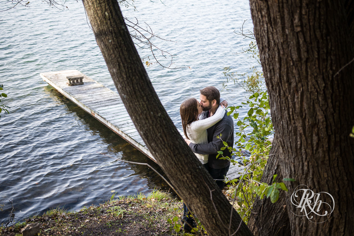 Man and woman kiss in front of lake during engagement photography session at home in Annandale, Minnesota.