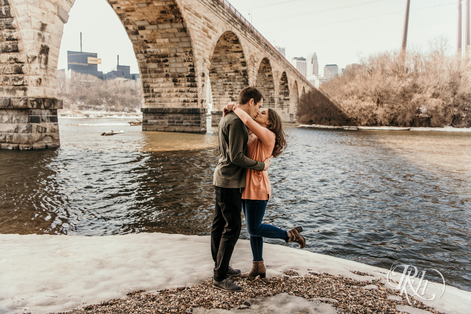 Man and women in sweater and jeans kiss in snow under Stone Arch Bridge in front of river in Minneapolis, Minnesota.