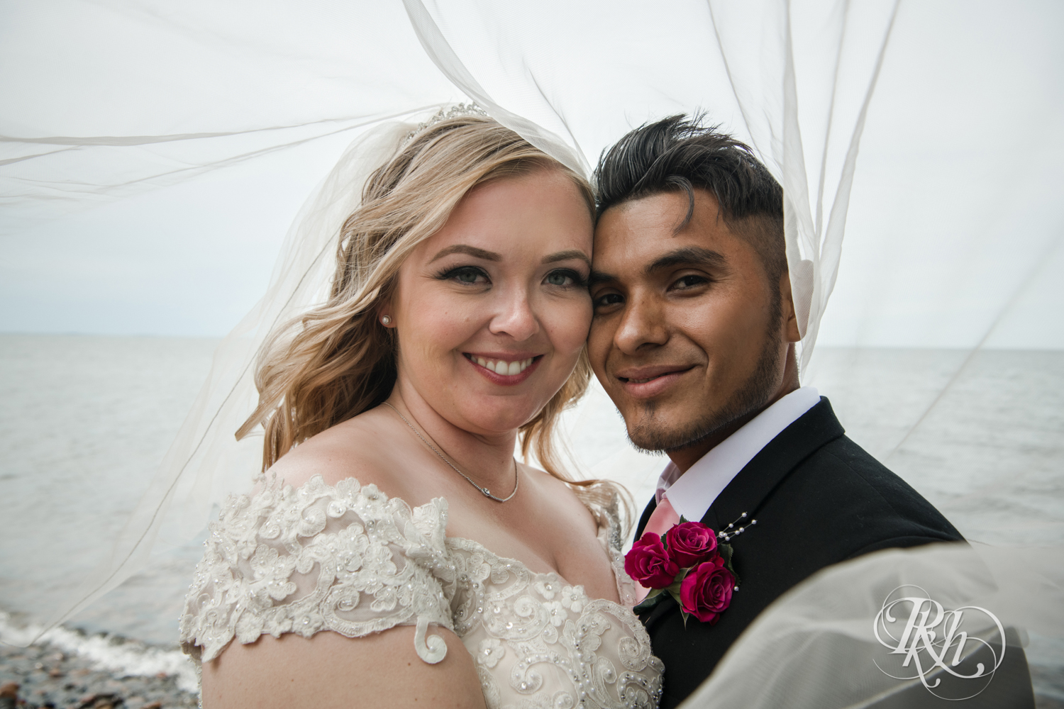 Bride and groom smile under veil in front of Lake Mille Lacs at Izatys Resort in Onamia, Minnesota.