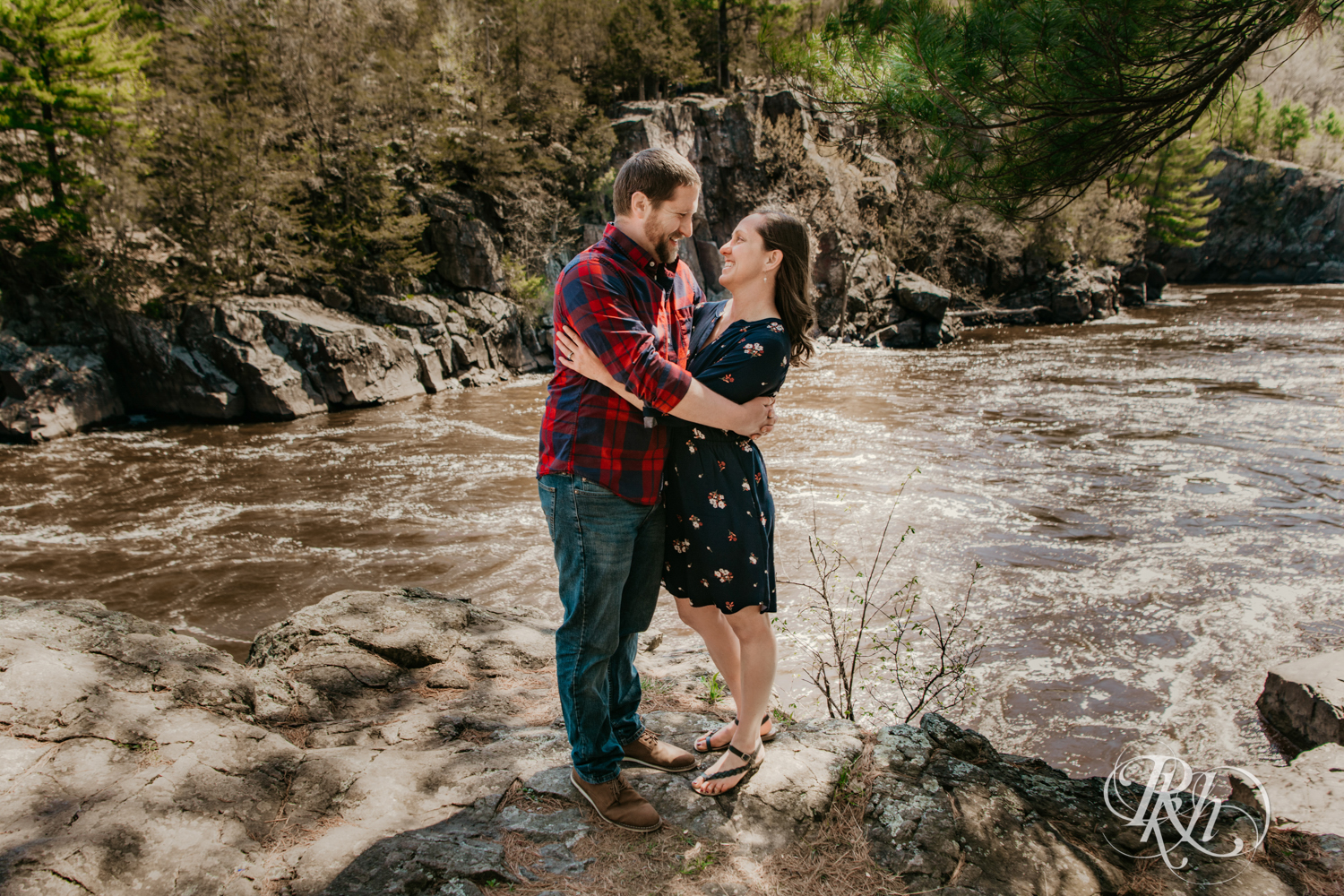 Man in flannel and jeans and woman in dress kiss on cliff over river in Taylor's Falls, Minnesota.