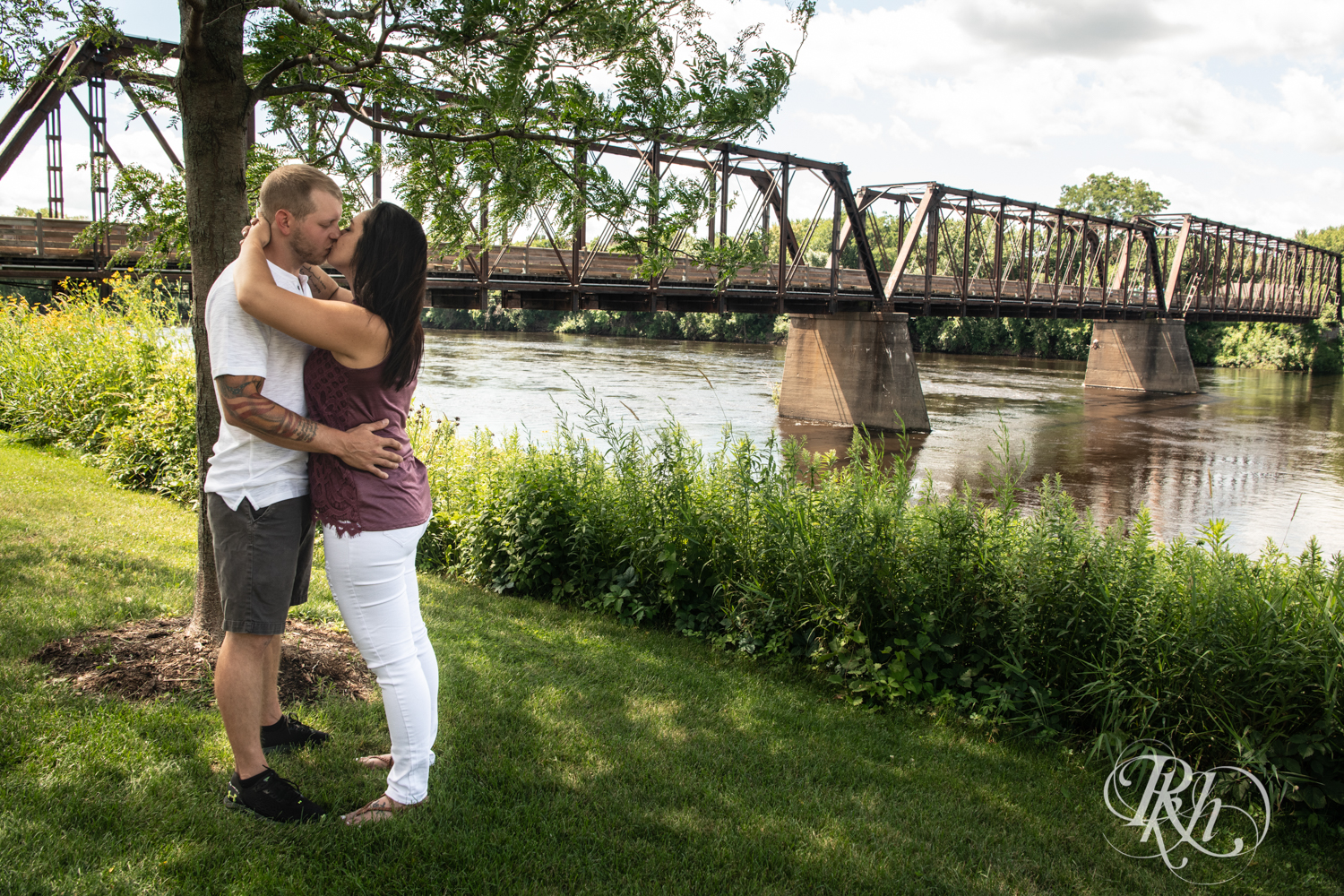 Man and woman smile in front of a bridge in Phoenix Park in Eau Claire, Wisconsin on a sunny day. 