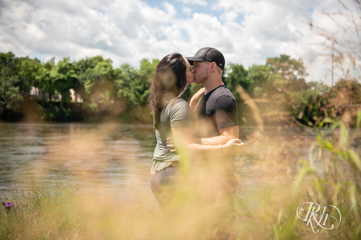 Man and woman kiss in front of a lake in Phoenix Park in Eau Claire, Wisconsin on a sunny day. 