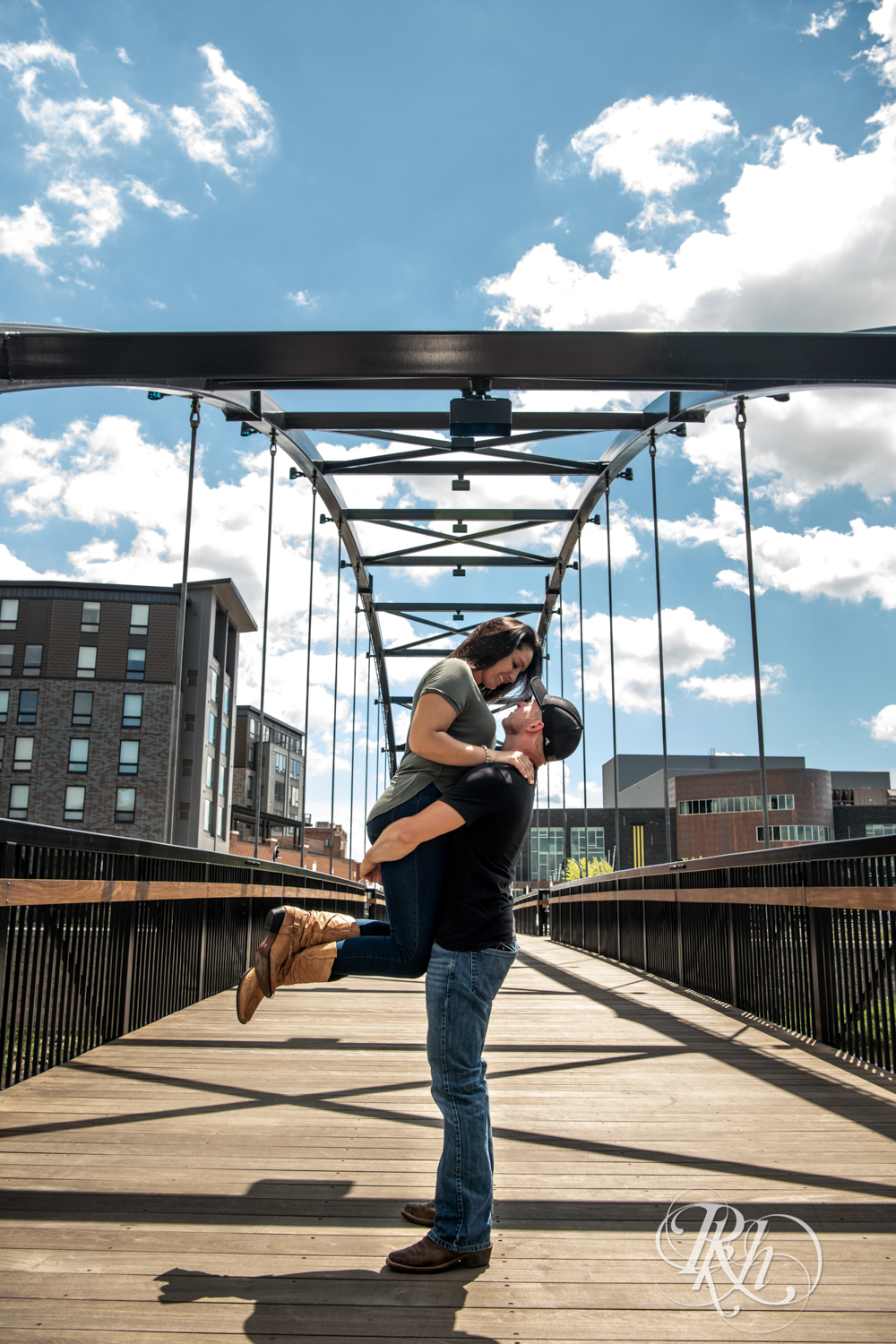 Man lifts woman in cowboy boots on a bridge in Phoenix Park in Eau Claire, Wisconsin on a sunny day. 