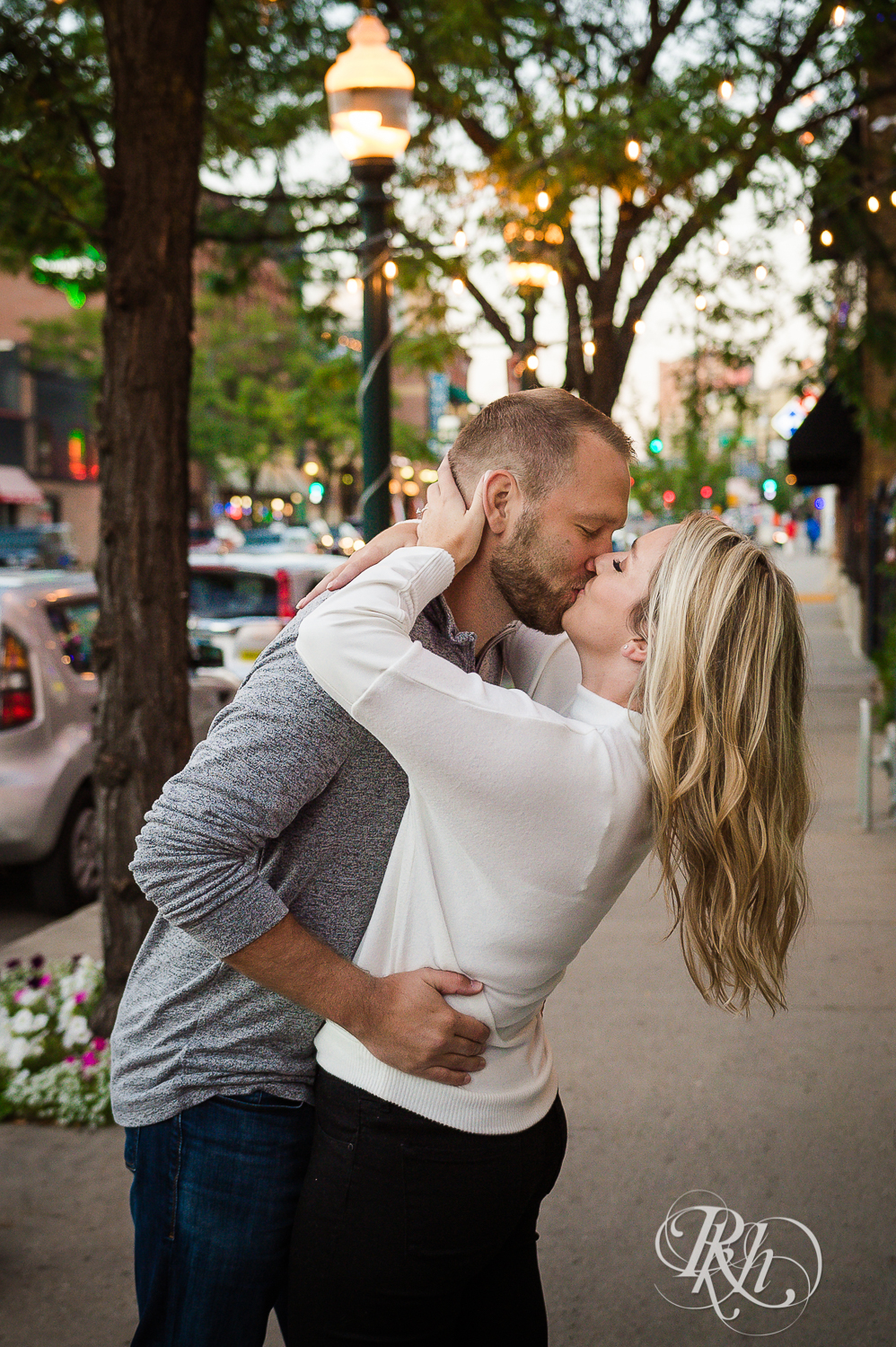 Blonde man and woman in sweaters and jeans kiss on city sidewalk at sunset in Saint Paul, Minnesota.