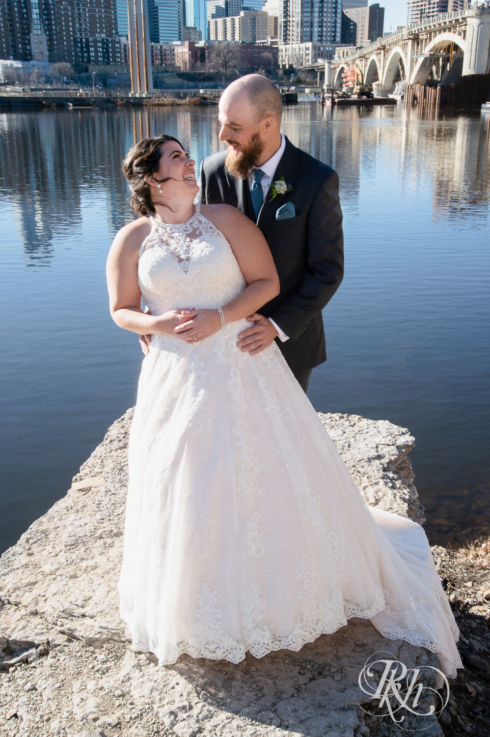 Bride and groom smile in front of river in Saint Anthony Main in Minneapolis, Minnesota.