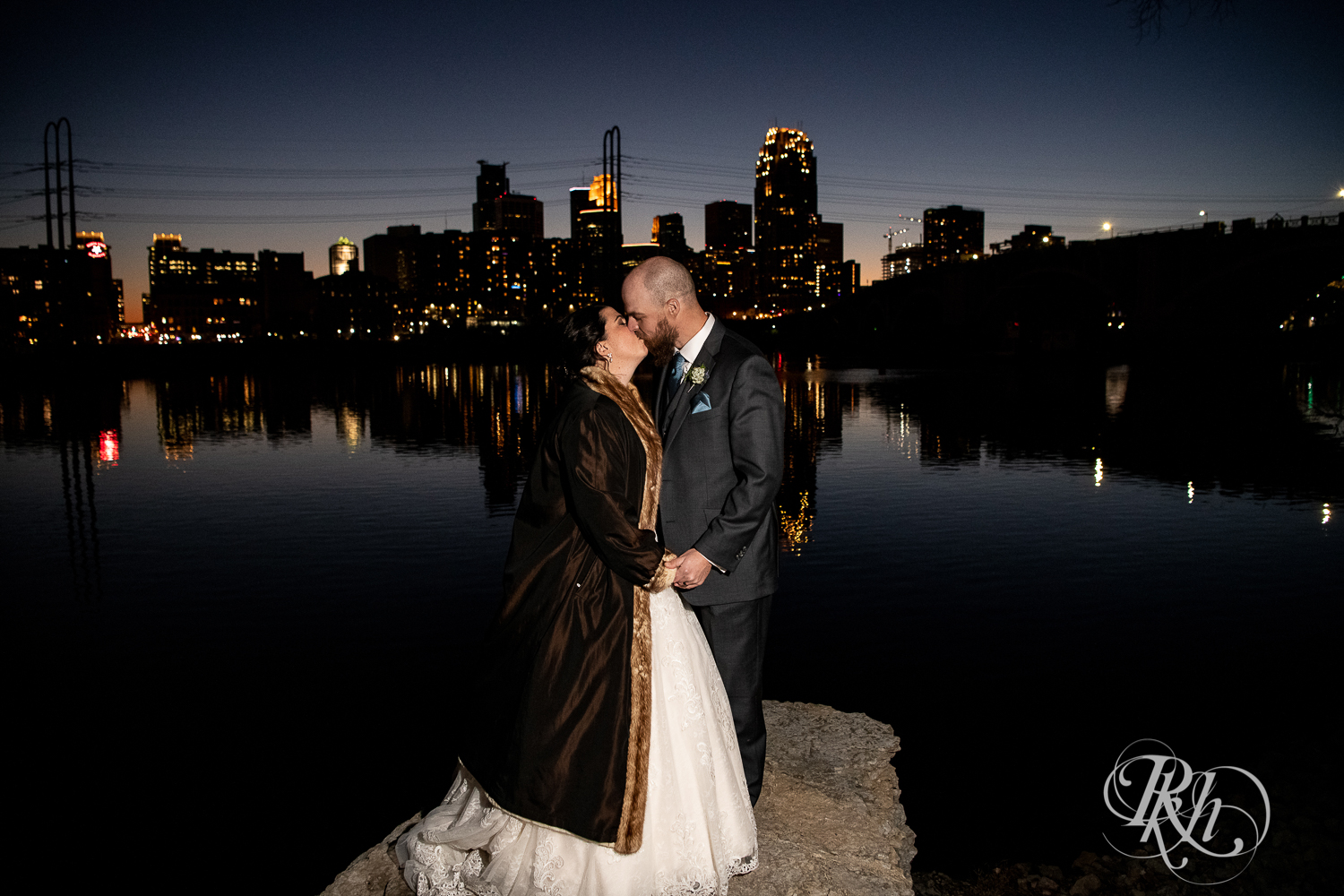 Bride and groom kiss at night in front of city skyline in Minneapolis, Minnesota.