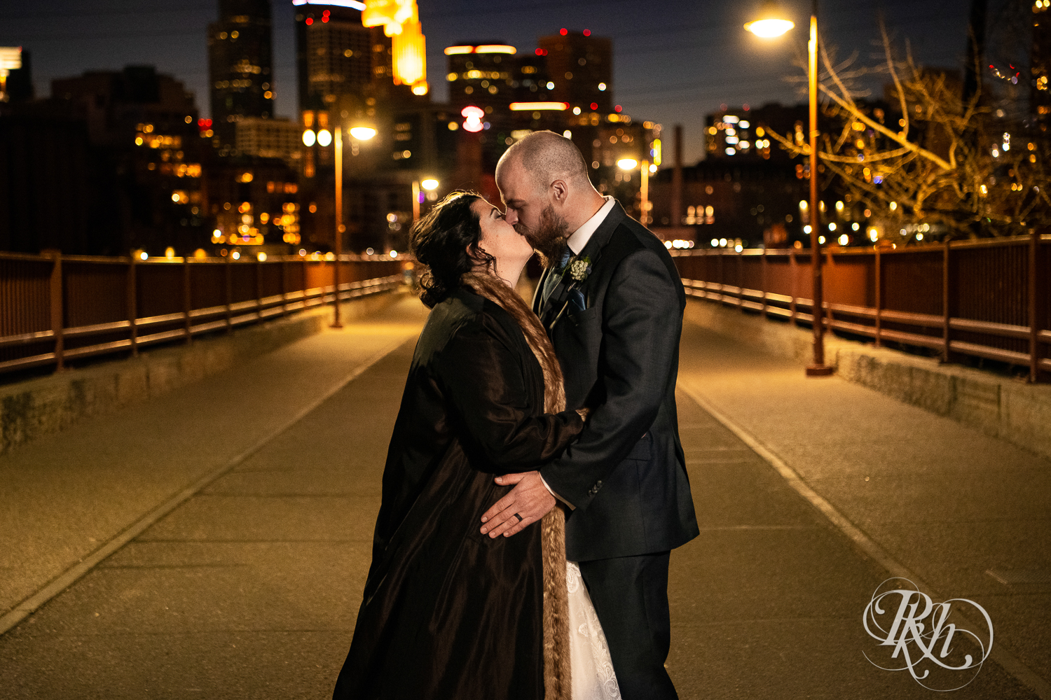 Bride and groom kiss at night in front of city skyline in Minneapolis, Minnesota on Stone Arch Bridge.