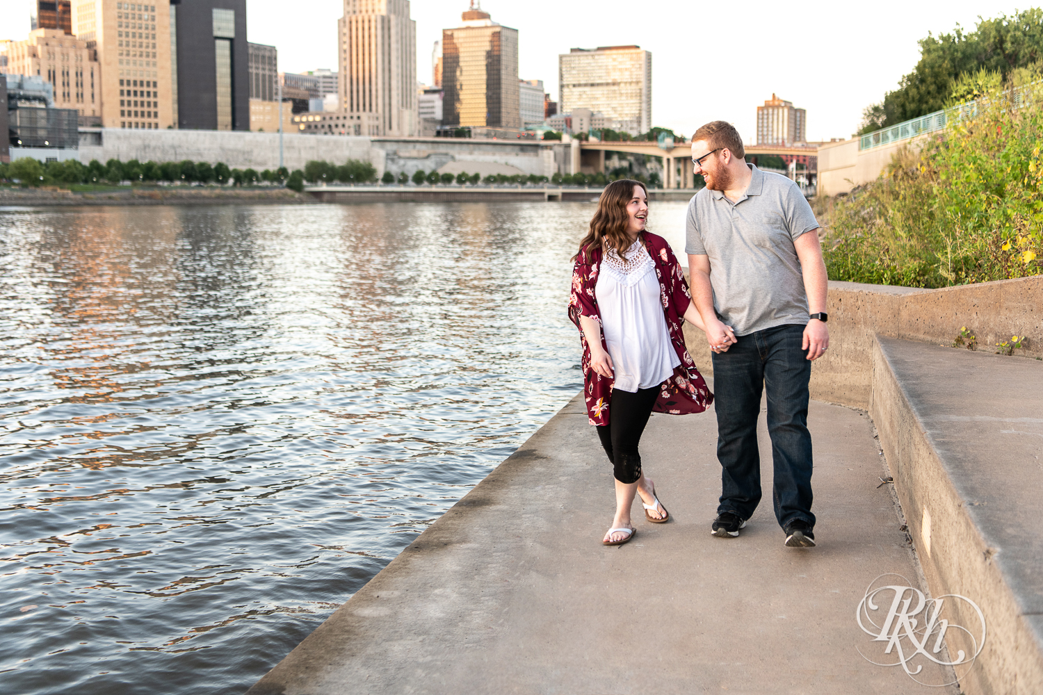 Man and woman walk together with city in background during golden hour on Harriet Island in Saint Paul, Minnesota.