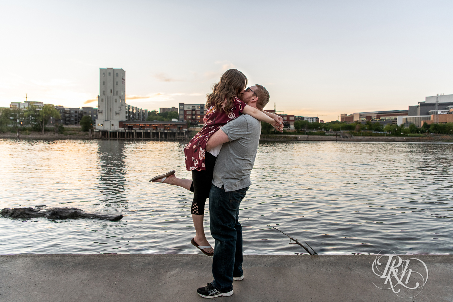Man lifts and kisses woman with city in background during golden hour on Harriet Island in Saint Paul, Minnesota.