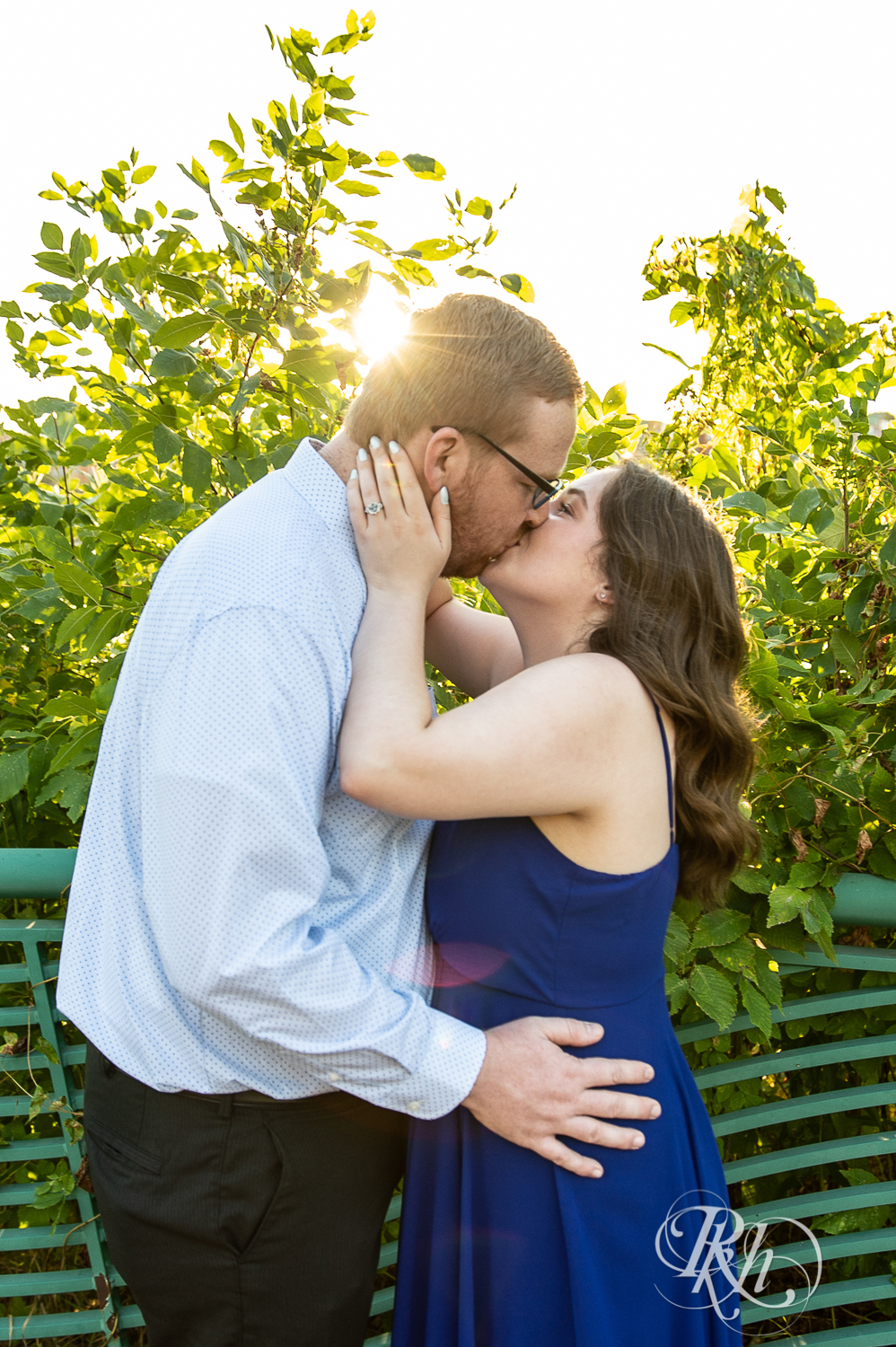 Man in glasses and woman in blue dress kiss during golden hour on Harriet Island in Saint Paul, Minnesota.