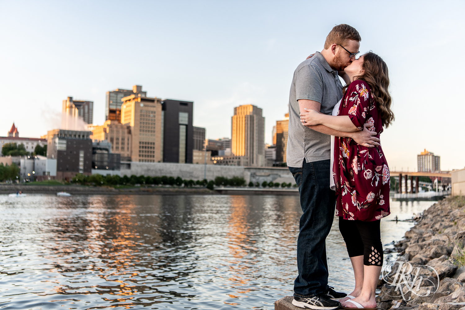 Man and woman kiss with city in background during golden hour on Harriet Island in Saint Paul, Minnesota.