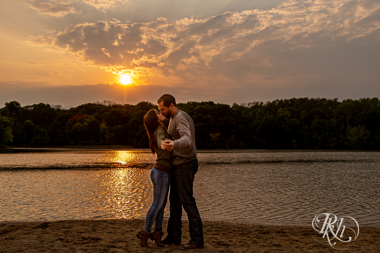 Man and woman in sweaters and jeans kiss in front of lake during sunset at Lebanon Hills Regional Park in Eagan, Minnesota.