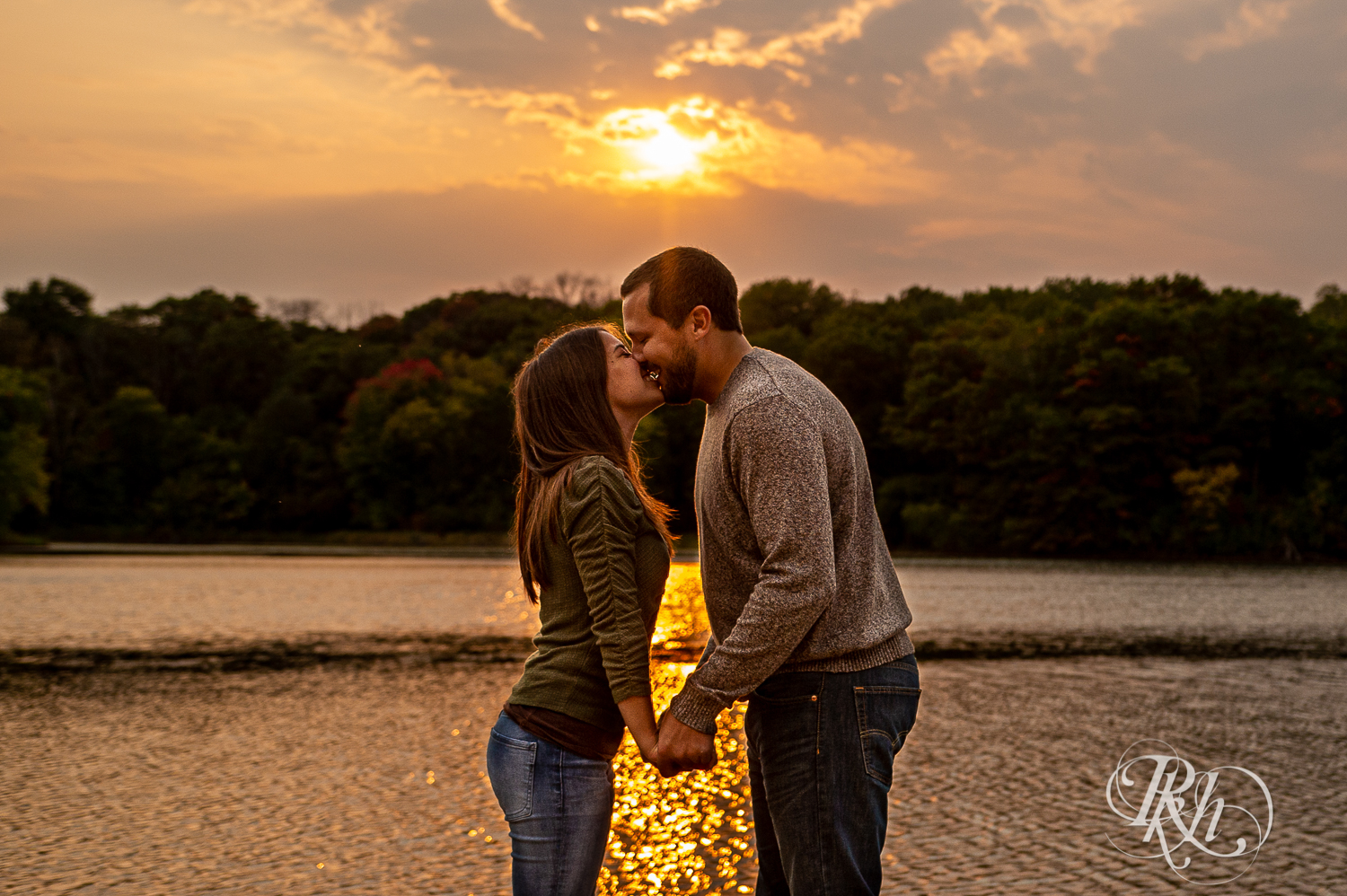 Man and woman in sweaters and jeans kiss in front of lake during sunset at Lebanon Hills Regional Park in Eagan, Minnesota.