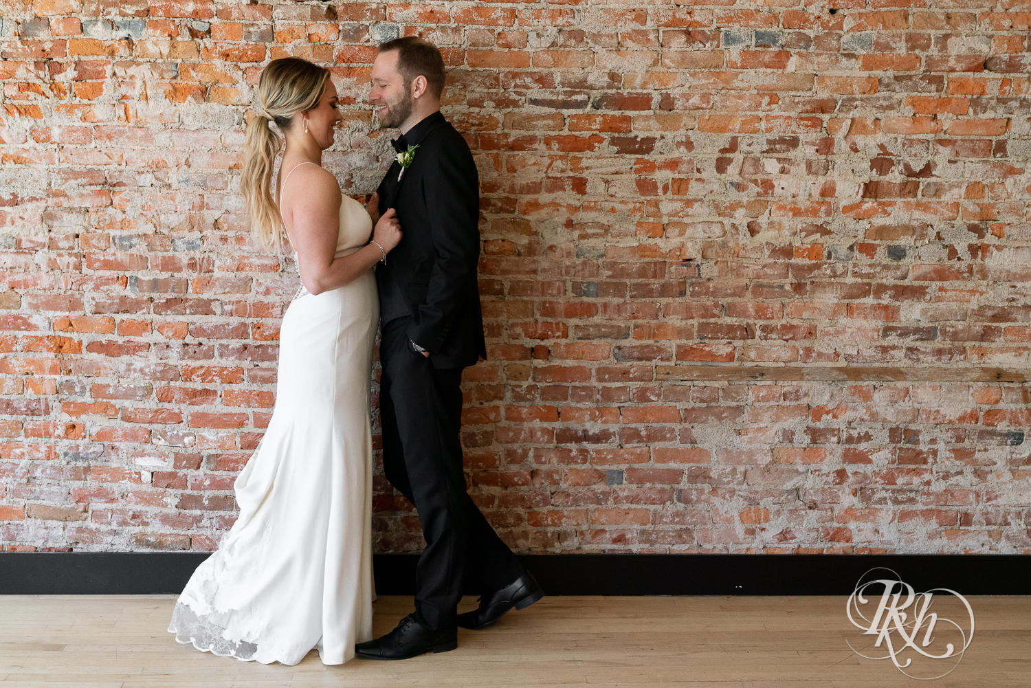 Bride and groom in black suit smile in front of brick wall in the 3 Ten Event Center in Faribault, Minnesota.