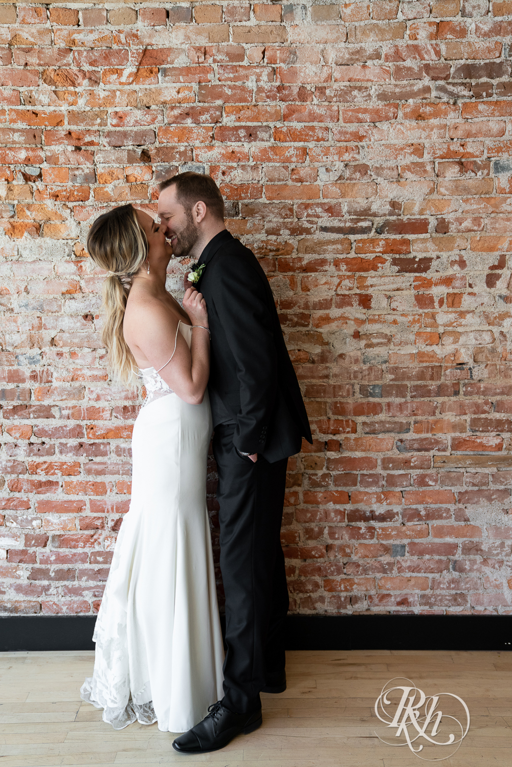 Bride and groom in black suit smile in front of brick wall in the 3 Ten Event Center in Faribault, Minnesota.