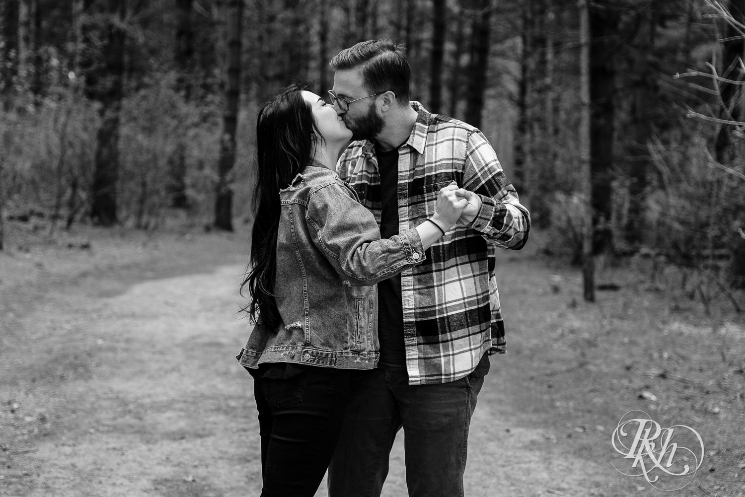 Man and woman in denim and flannel kiss between trees at Lebanon Hills Regional Park in Eagan, Minnesota. 