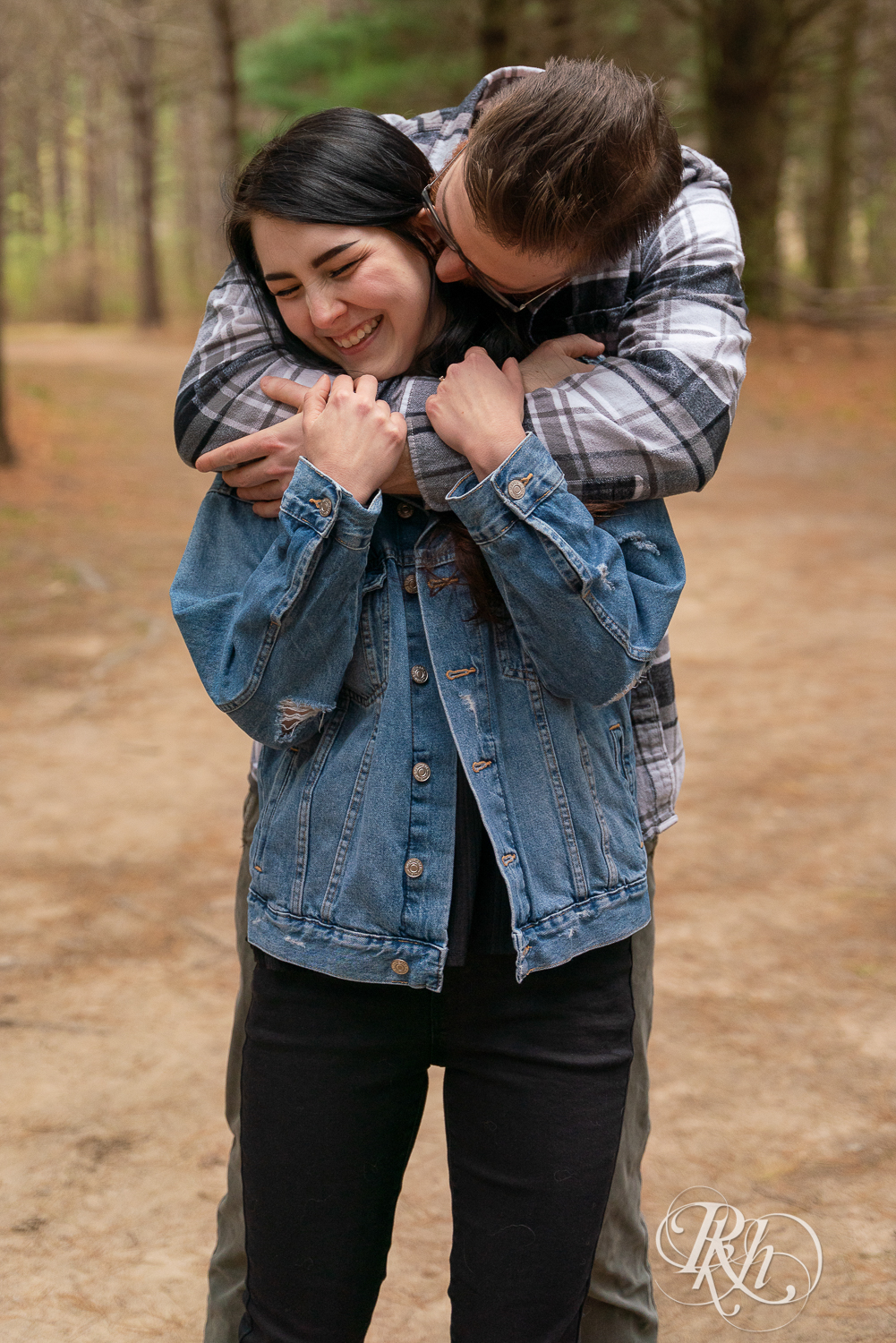 Man and woman in denim and flannel snuggle between trees at Lebanon Hills Regional Park in Eagan, Minnesota. 