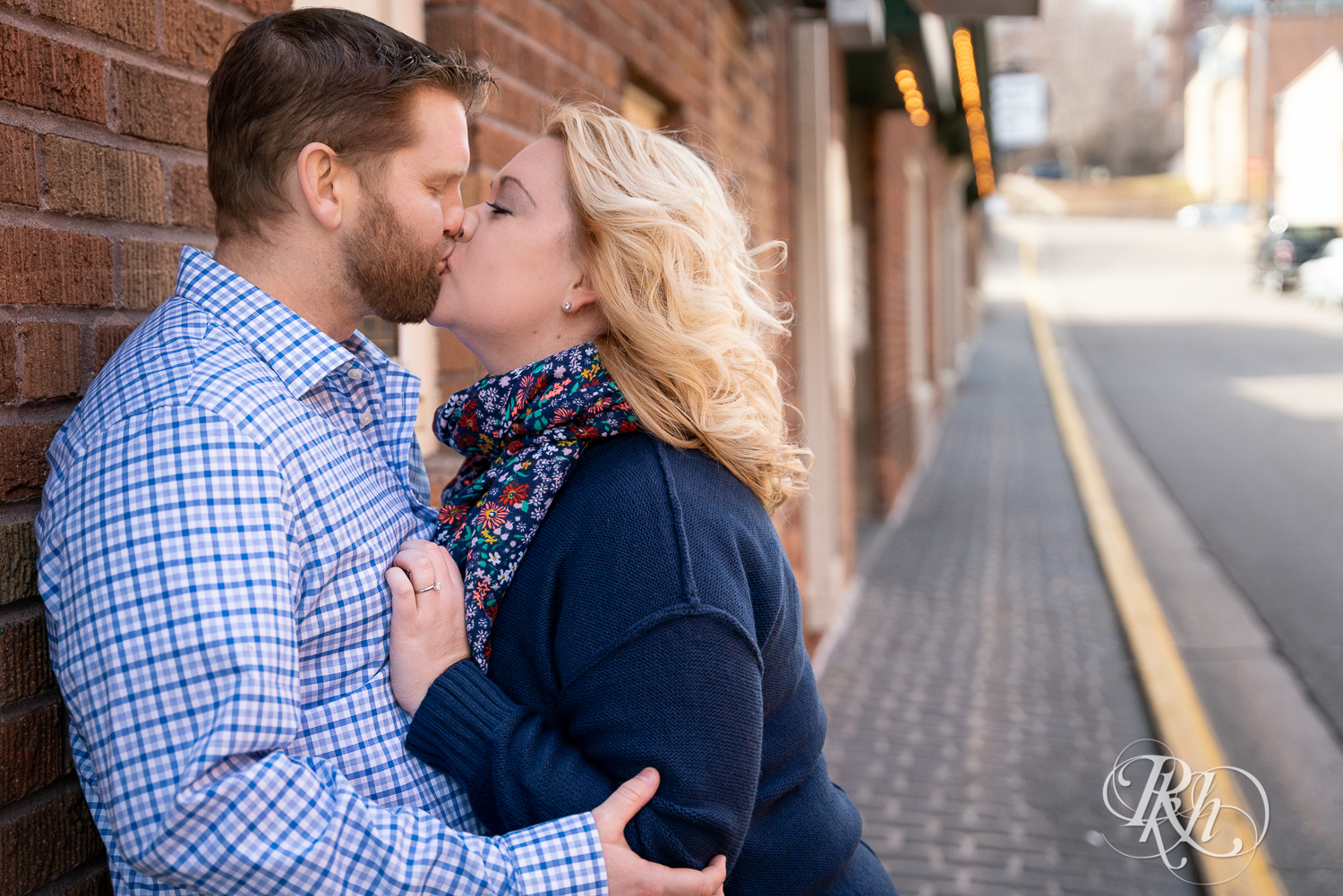 Blond woman and bearded man kiss against brick wall in Stillwater, Minnesota.