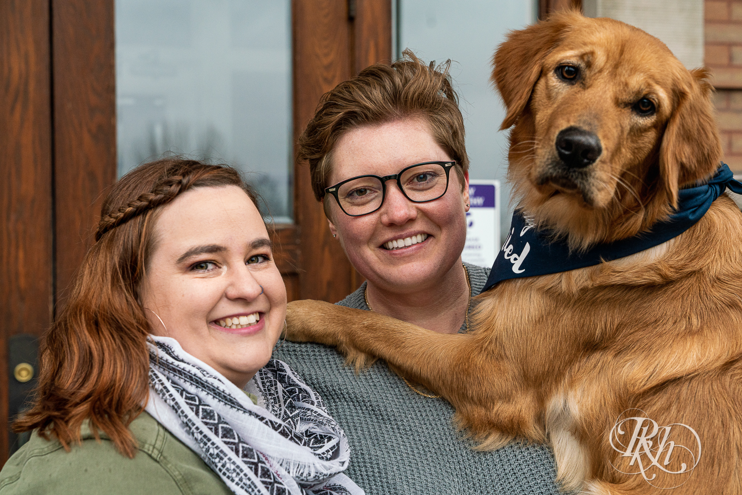 Lesbian couple smile with their Golden Retriever in front of Saint Catherine's in Saint Paul, Minnesota. 
