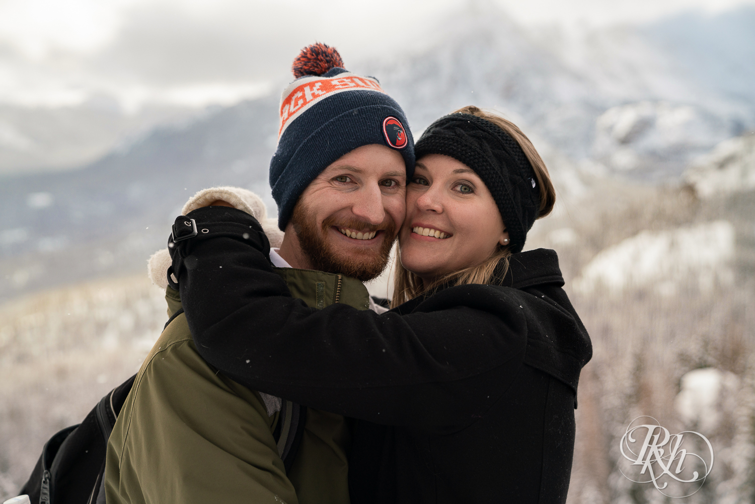 Man and woman in winter jackets and hats smile in front of Rocky Mountains view in Colorado.