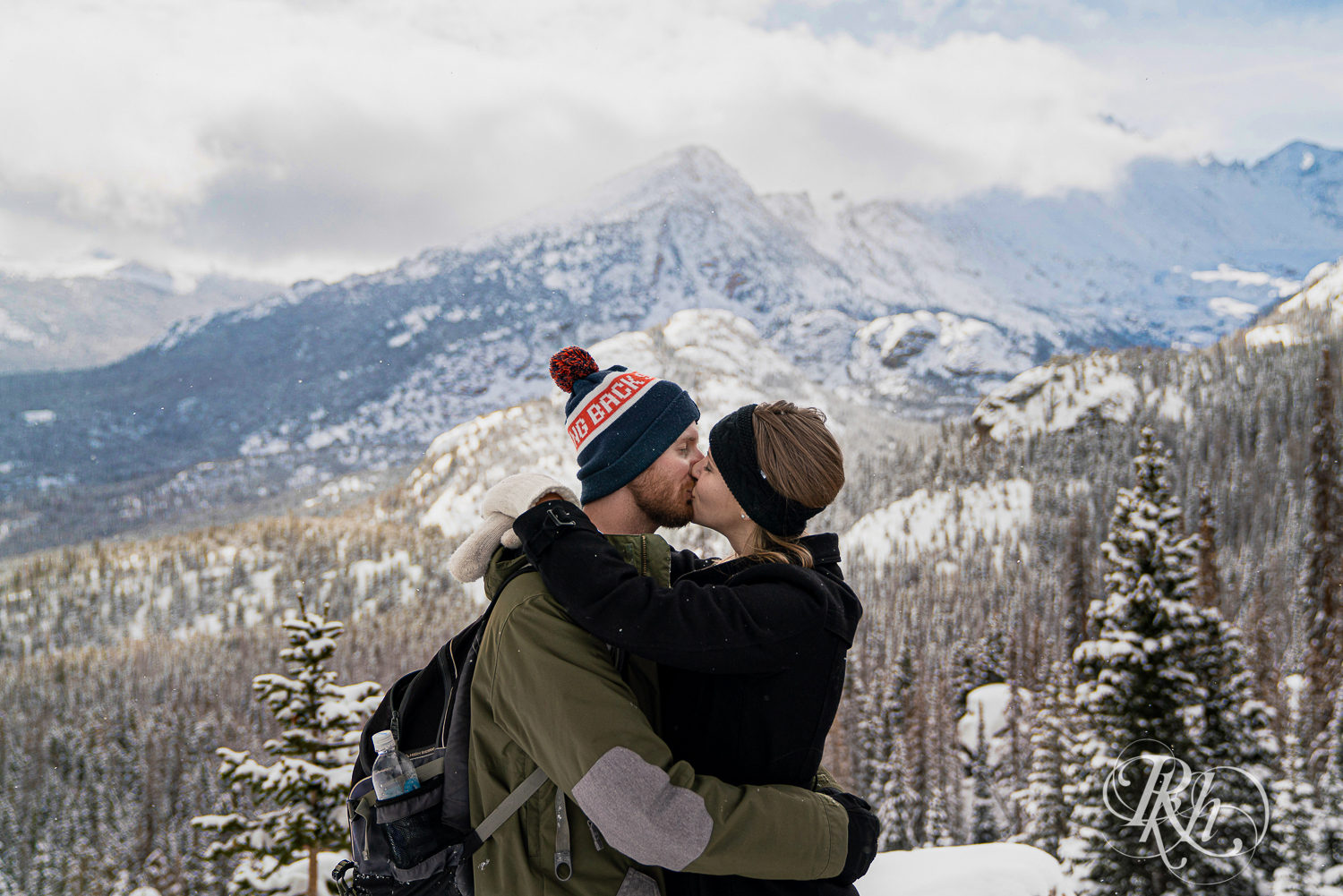 Man and woman in winter jackets and hats kiss in front of Rocky Mountains view in Colorado.