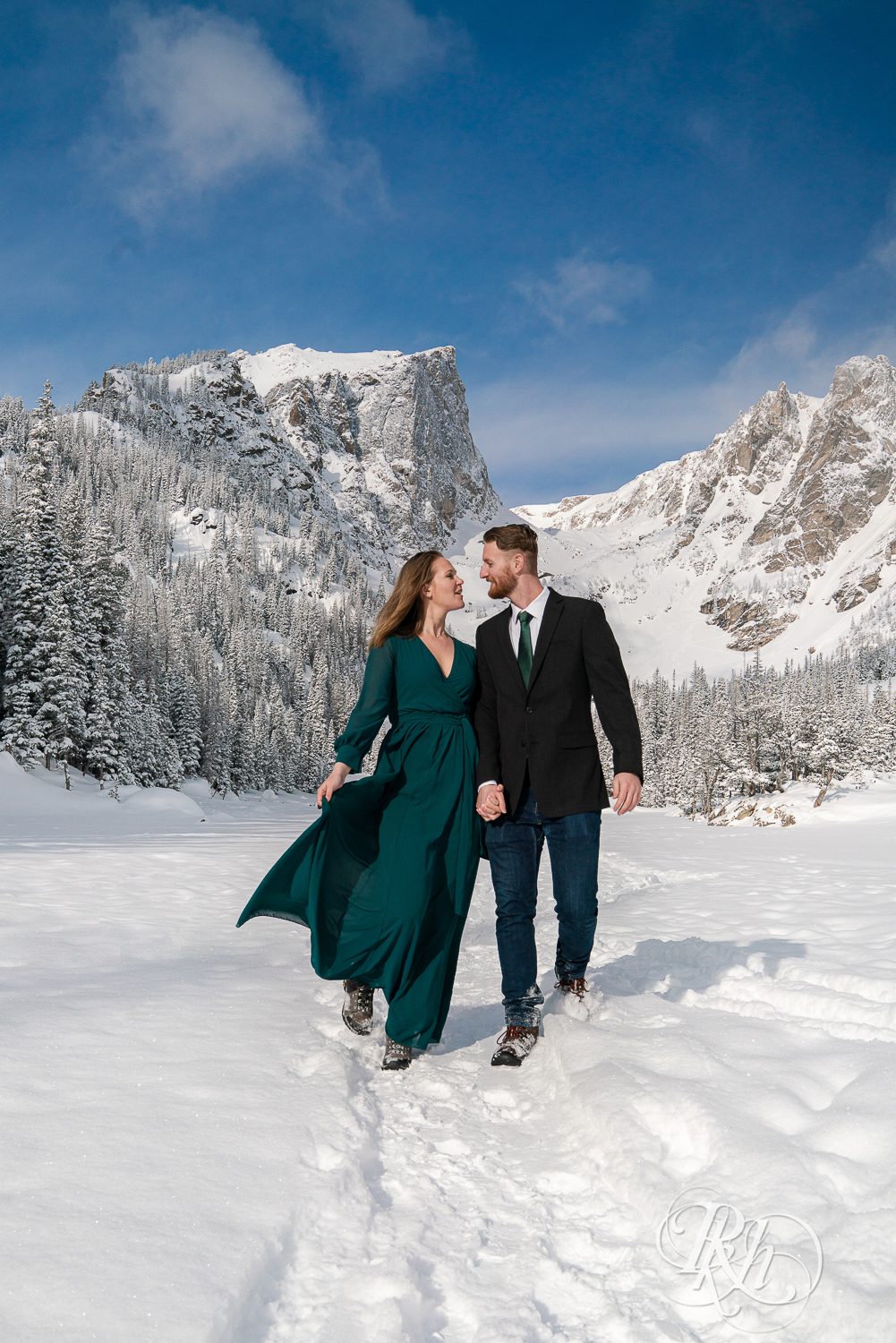 Man in suit and woman in green dress walk in snow on Dream Lake in Rocky Mountain National Park, Colorado.