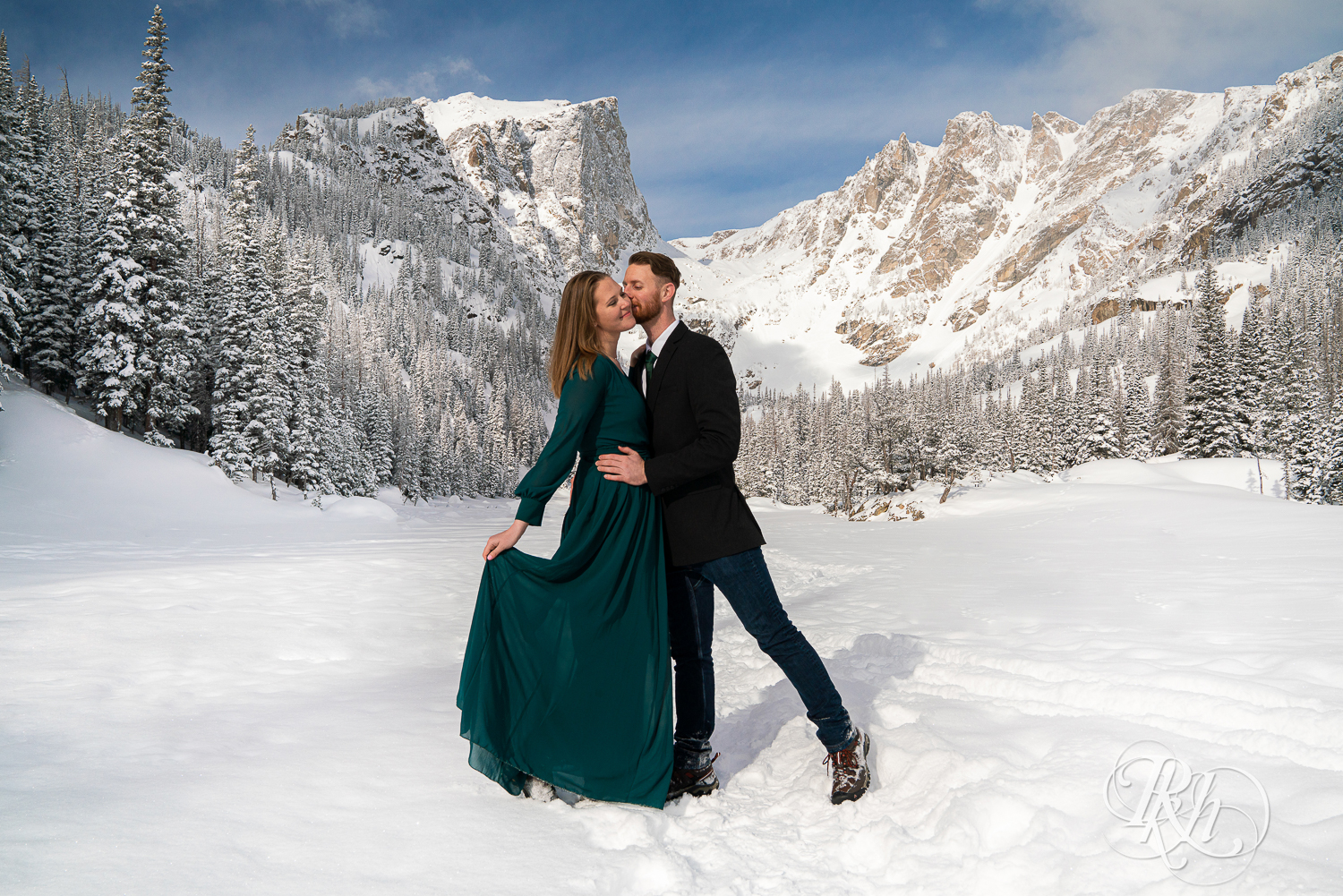 Man in suit and woman in green dress kiss in snow on Dream Lake in Rocky Mountain National Park, Colorado.