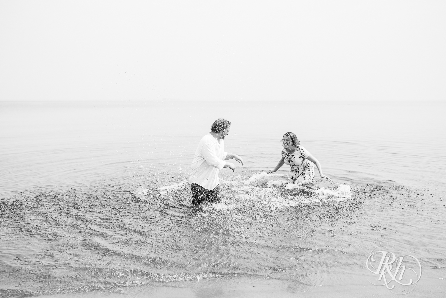 Man and woman splash water at each other in Lake Superior during engagement photos on Park Point Beach in Duluth, Minnesota.