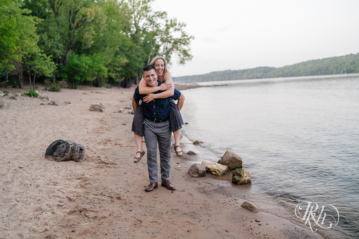 Man carries blonde woman in blue dress on his back on beach at Afton State Park in Hastings, Minnesota.