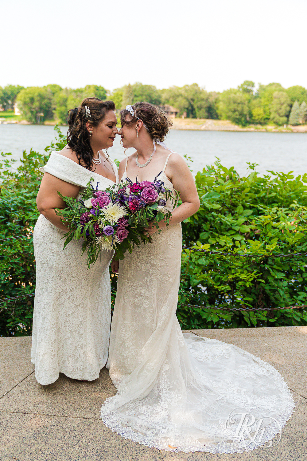 Lesbian brides holding flowers and smiling by the water at wedding in Champlin, Minnesota.