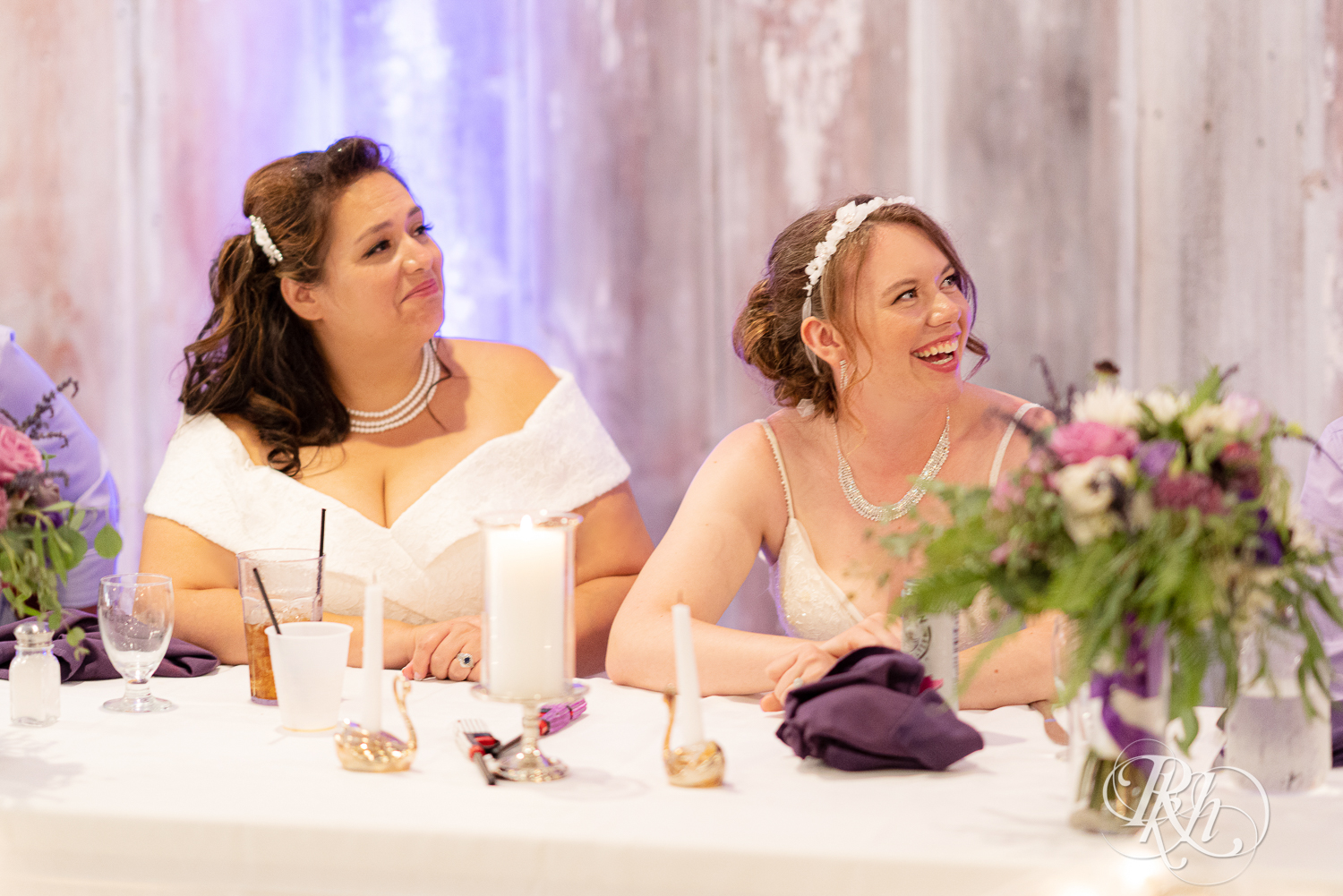 Lesbian brides smile at head table at wedding reception at Willy McCoy's in Champlin, Minnesota.