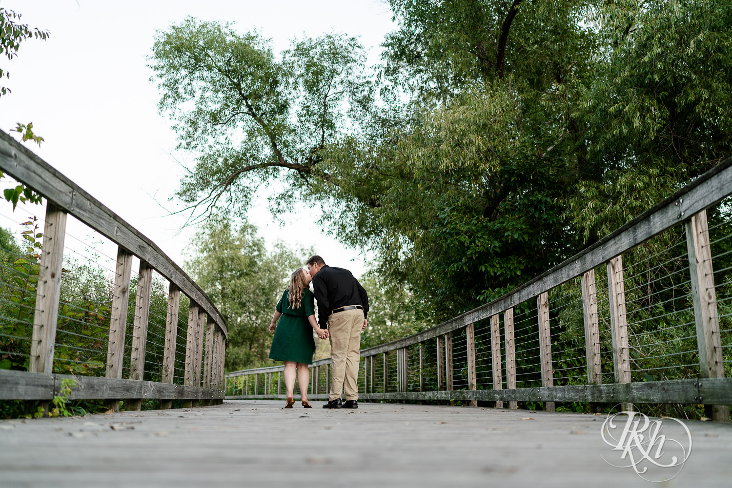 Man in black dress shirt walking with and kissing woman in green dress on a bridge in Eagan, Minnesota.