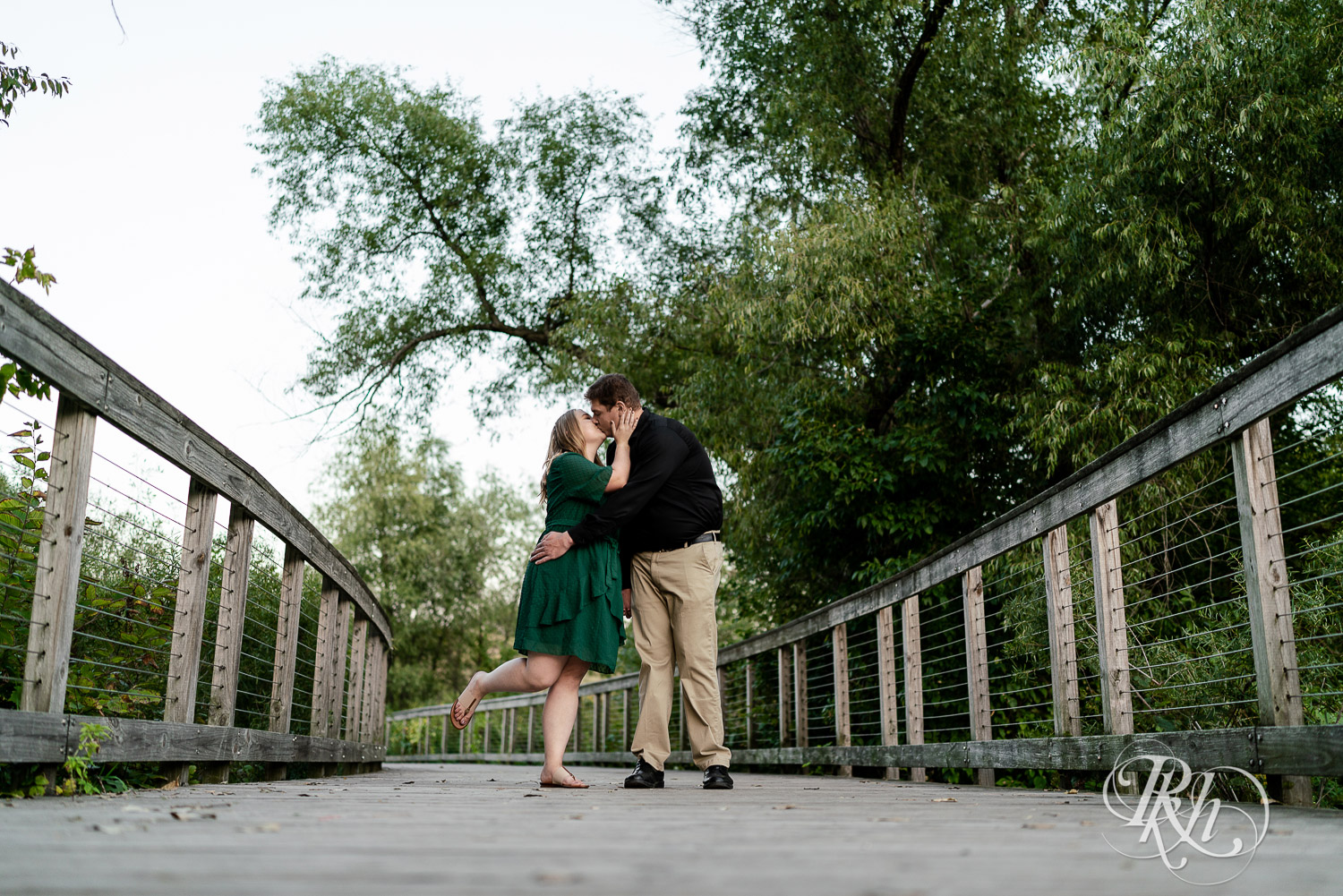 Man in black dress shirt walking with and kissing woman in green dress on a bridge in Eagan, Minnesota.